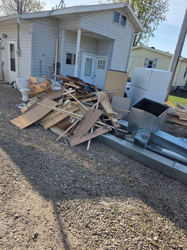 Piled demolition debris beside a small white house with a gravel yard and parked equipment.
