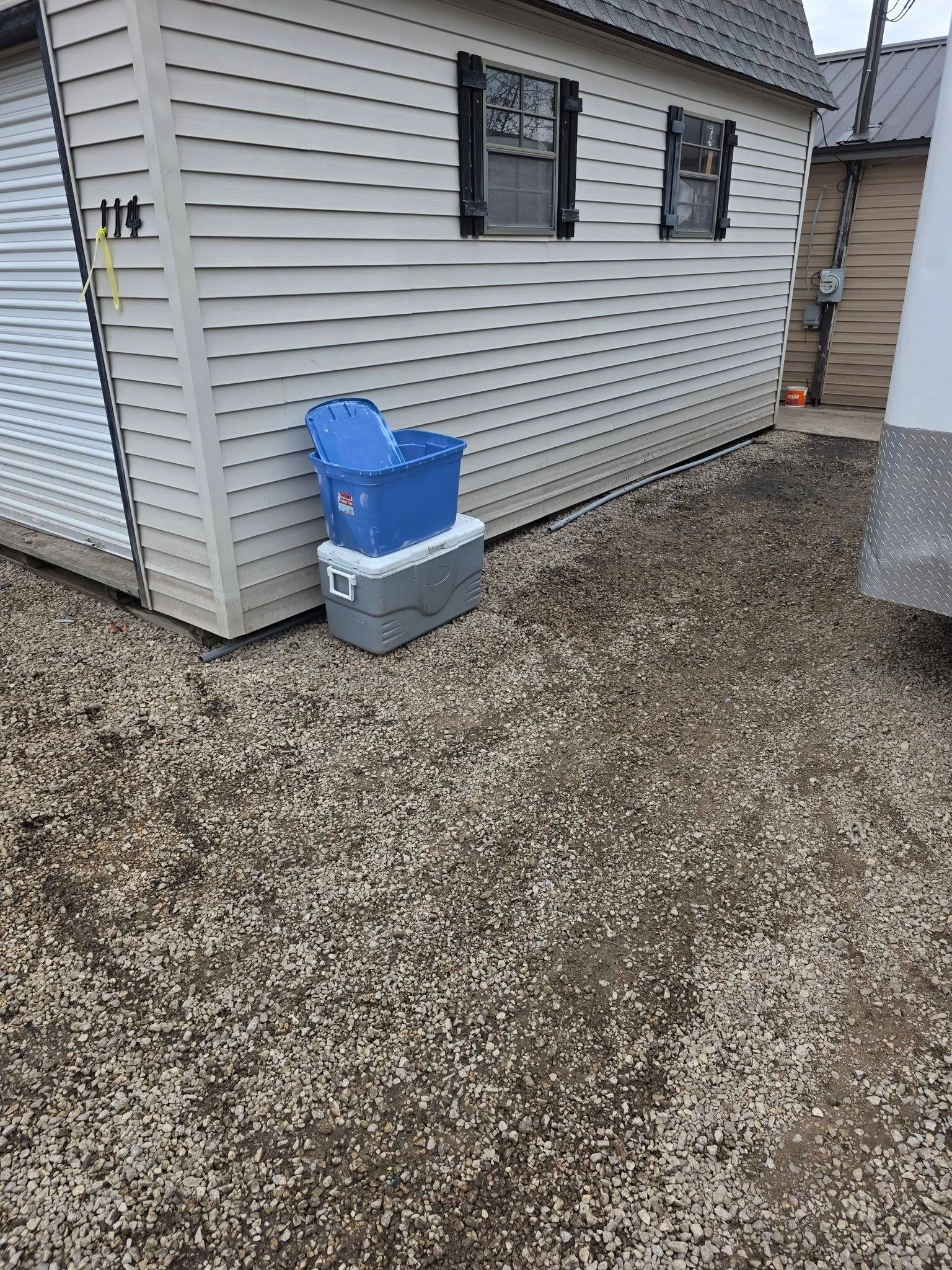 Gravel yard beside a white-sided house with two windows and a blue bin on a gray container.
