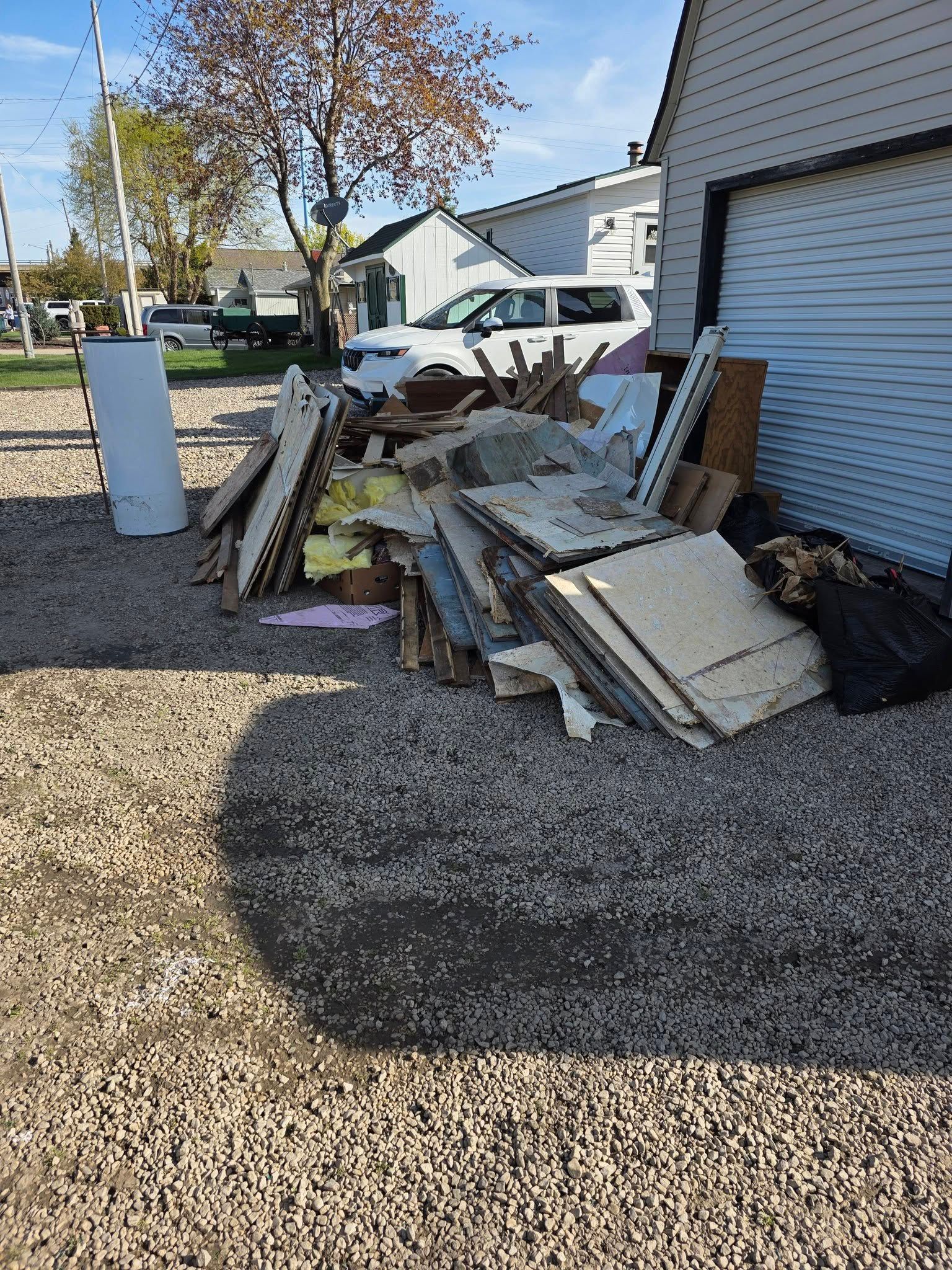 Piled construction debris and scrap wood beside a white garage in a gravel driveway