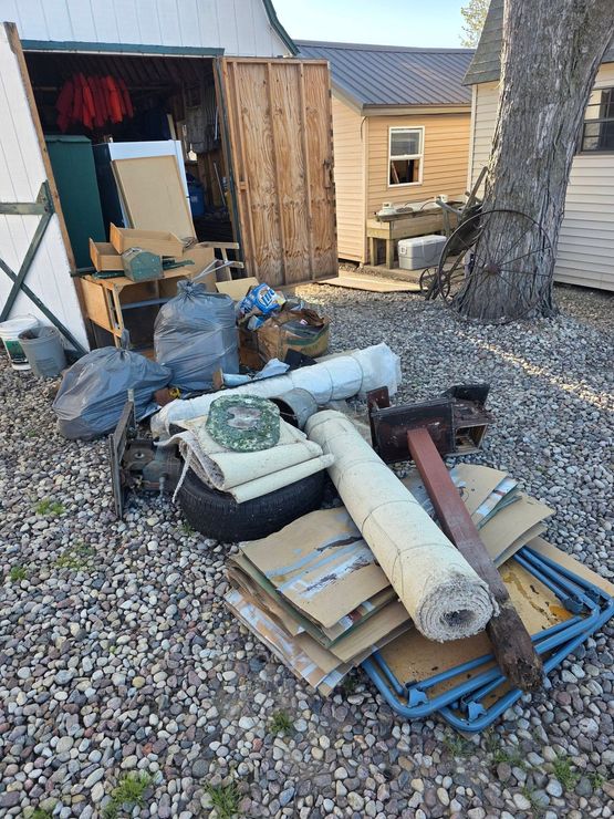 Cluttered yard with stacked scrap materials, rolled insulation, pallets, and debris beside a shed and tree trunk