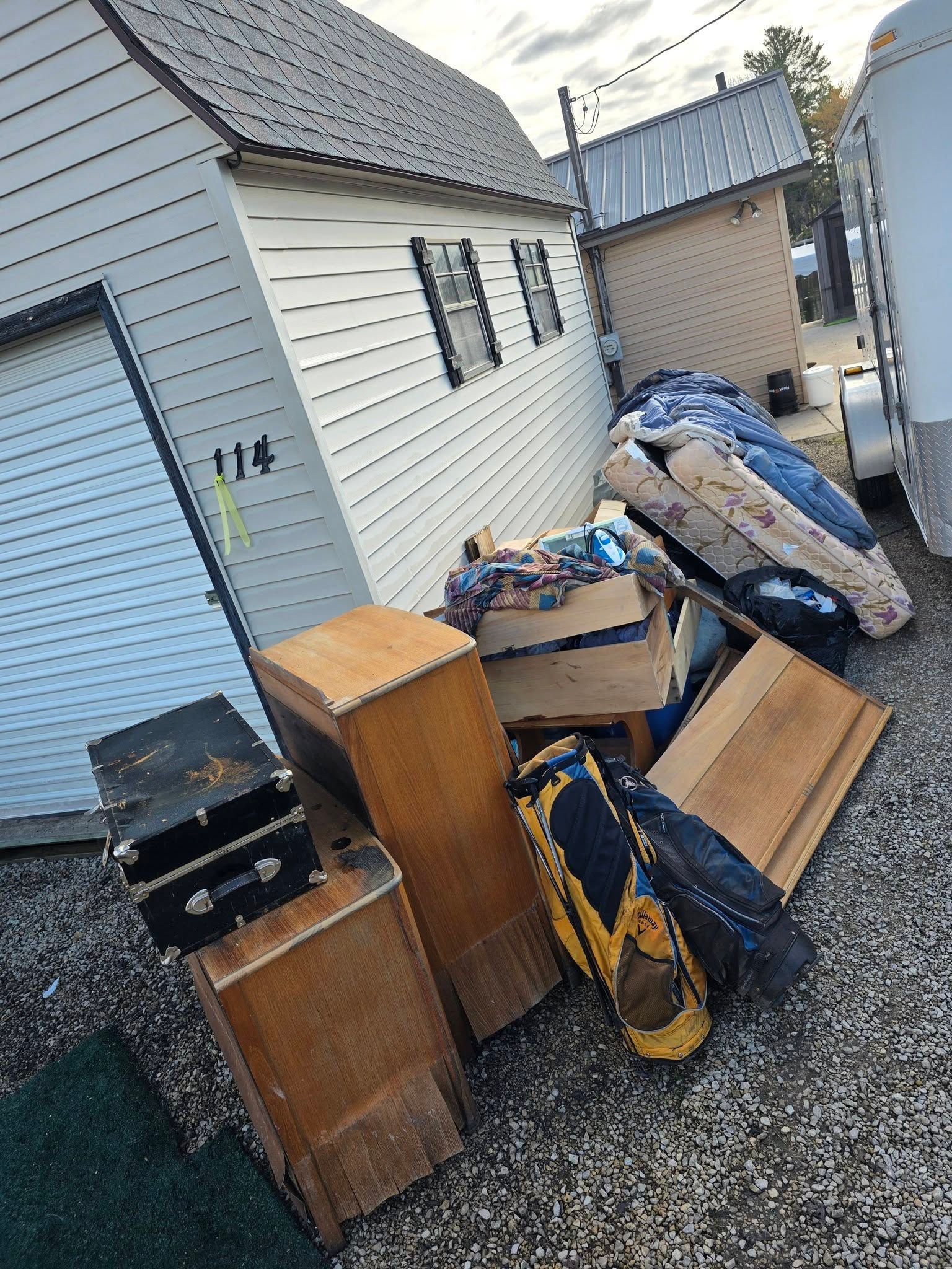 Outdoor alley beside white sheds with a pile of broken furniture, boxes, and trash bags.