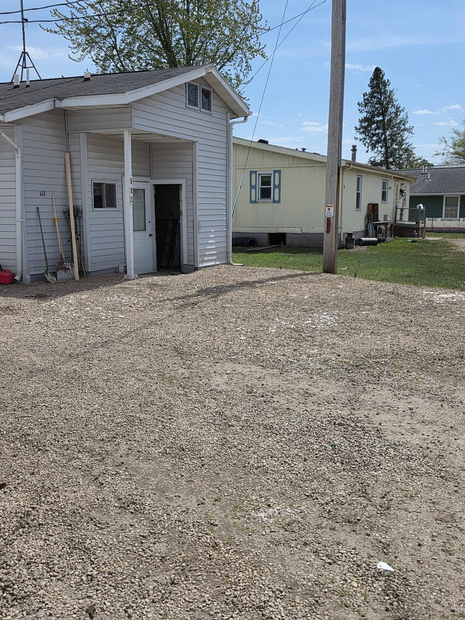 Small white shed-like building with open doors beside a beige house on a gravel lot under a clear sky
