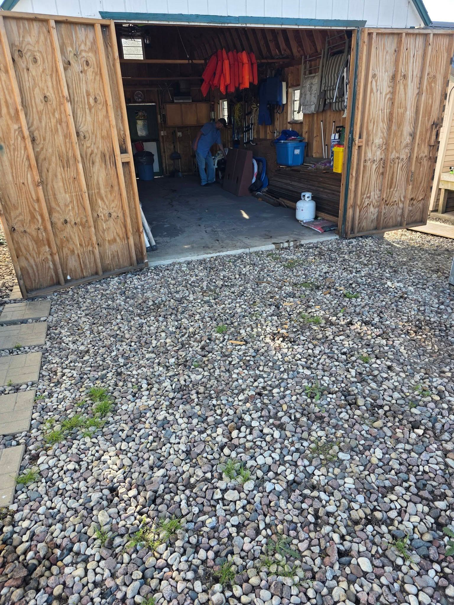 Open wooden garage with tools and hanging red jackets, gravel driveway in front.