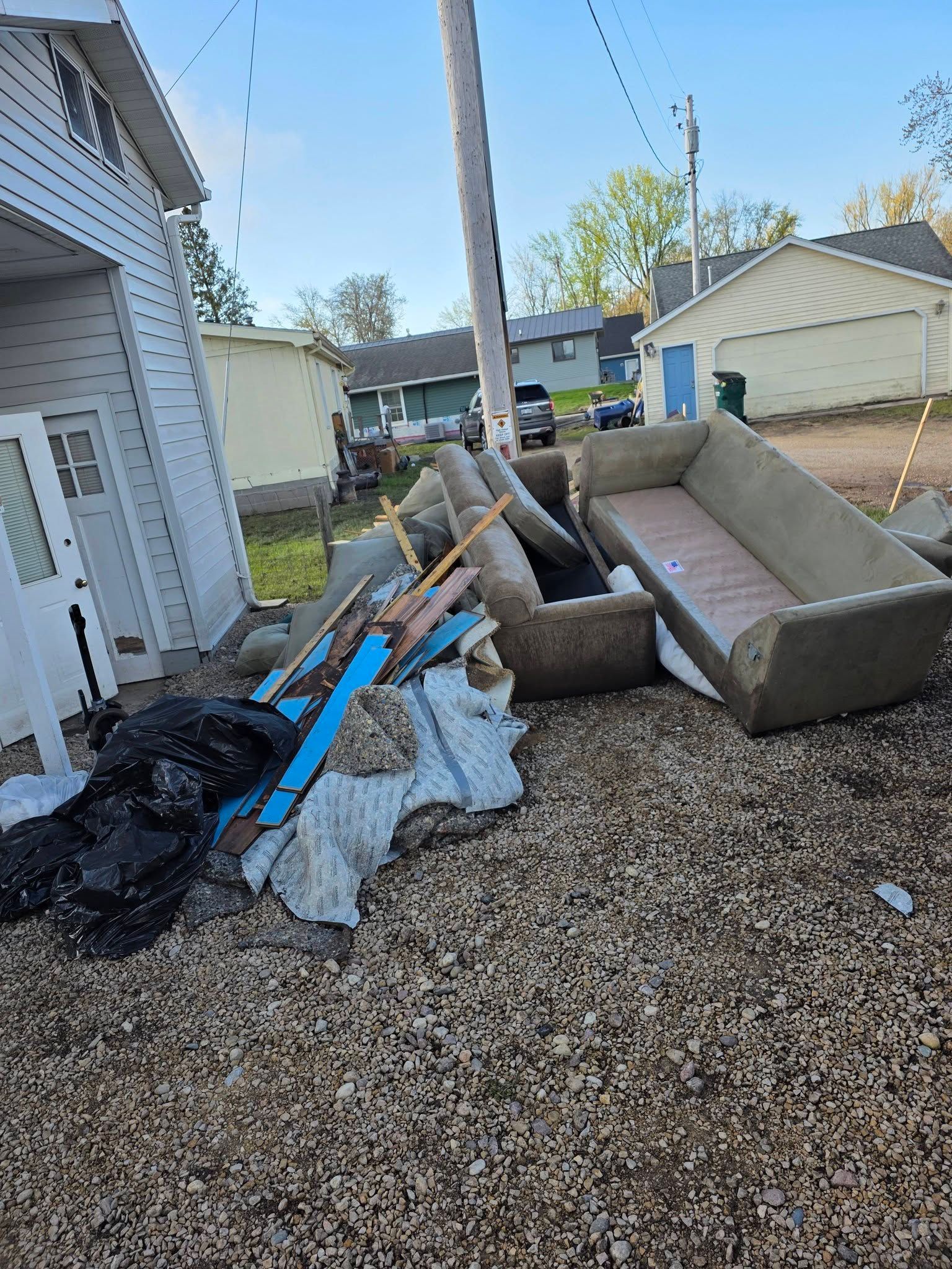 Storm debris piled beside a house, with broken furniture, branches, and tarps on gravel.