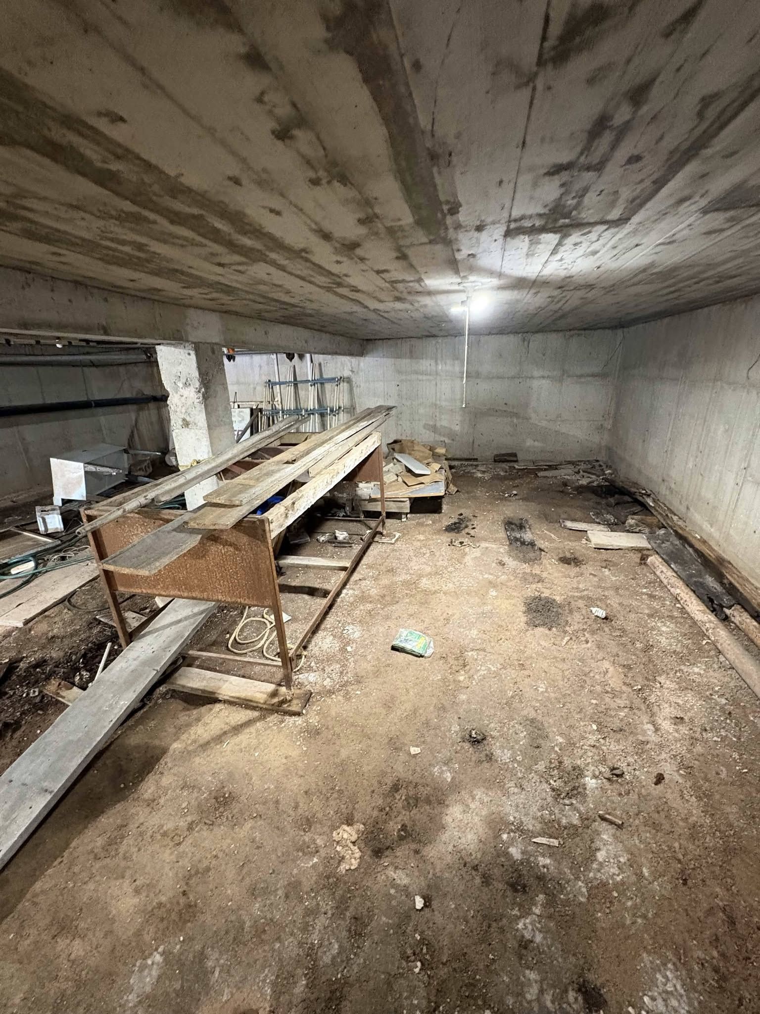 Abandoned basement room with peeling ceiling, long wooden bench, and debris on a dirty concrete floor.