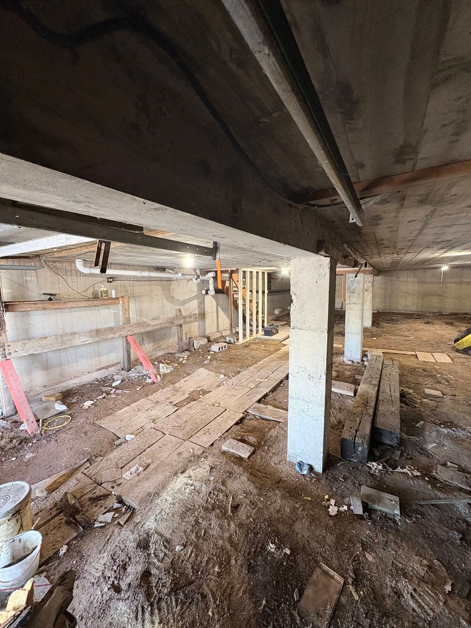 Unfinished basement with exposed beams, concrete columns, dirt floor, and construction materials.