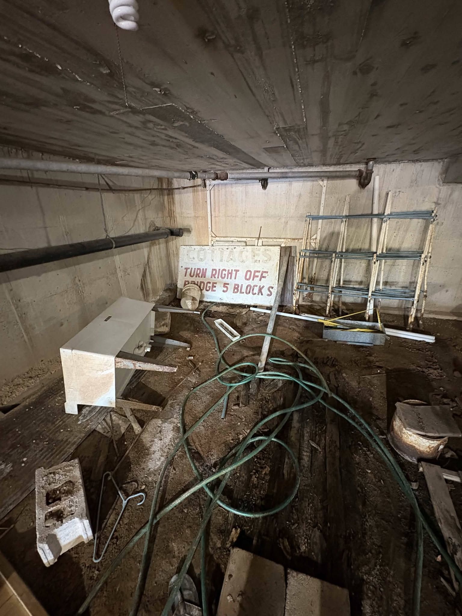 Damp, cluttered basement with pipes, a workbench, coiled hoses, and a sign on the back wall