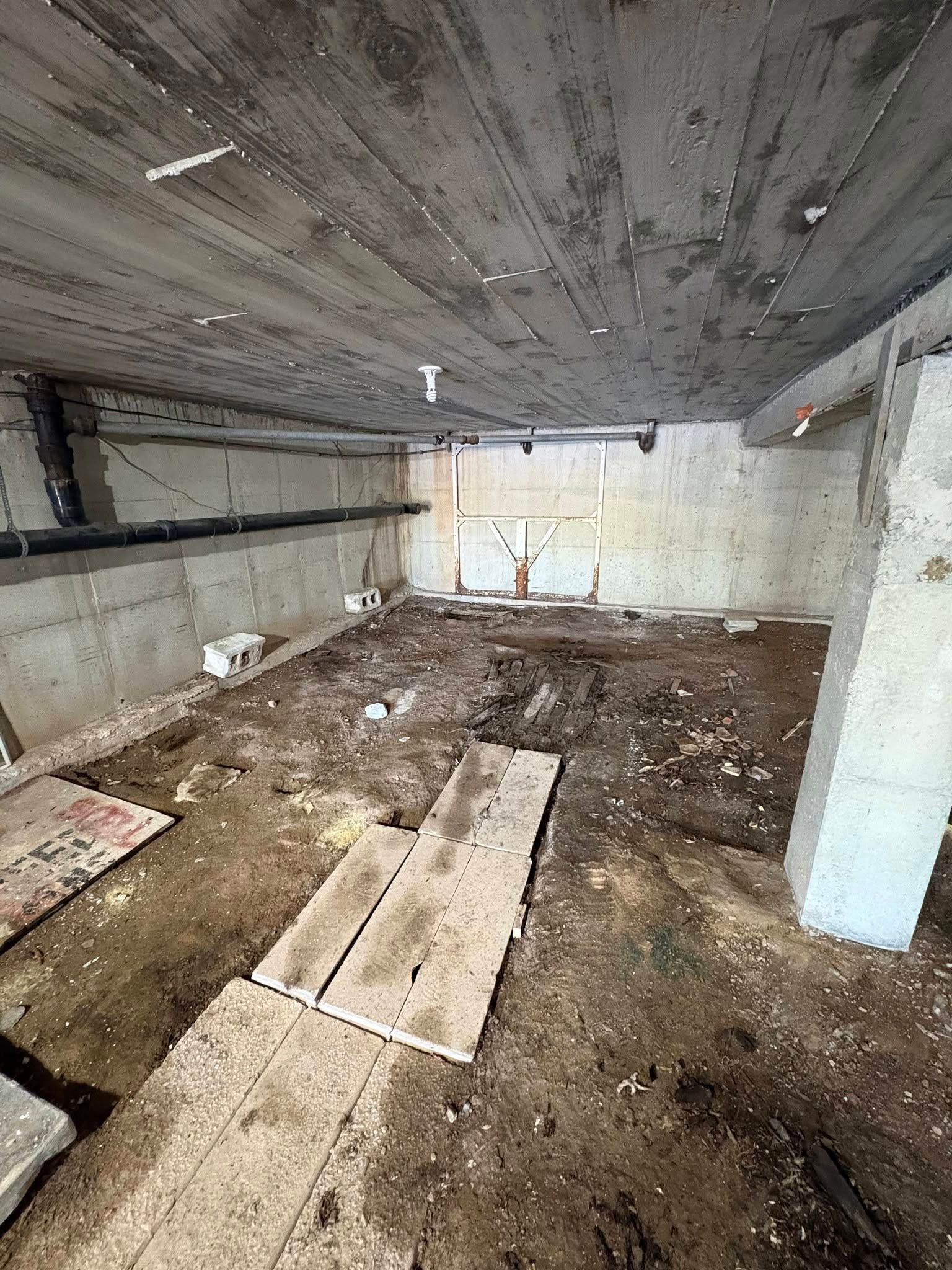 Abandoned unfinished room with dirt floor, exposed concrete ceiling, and scattered wooden planks.