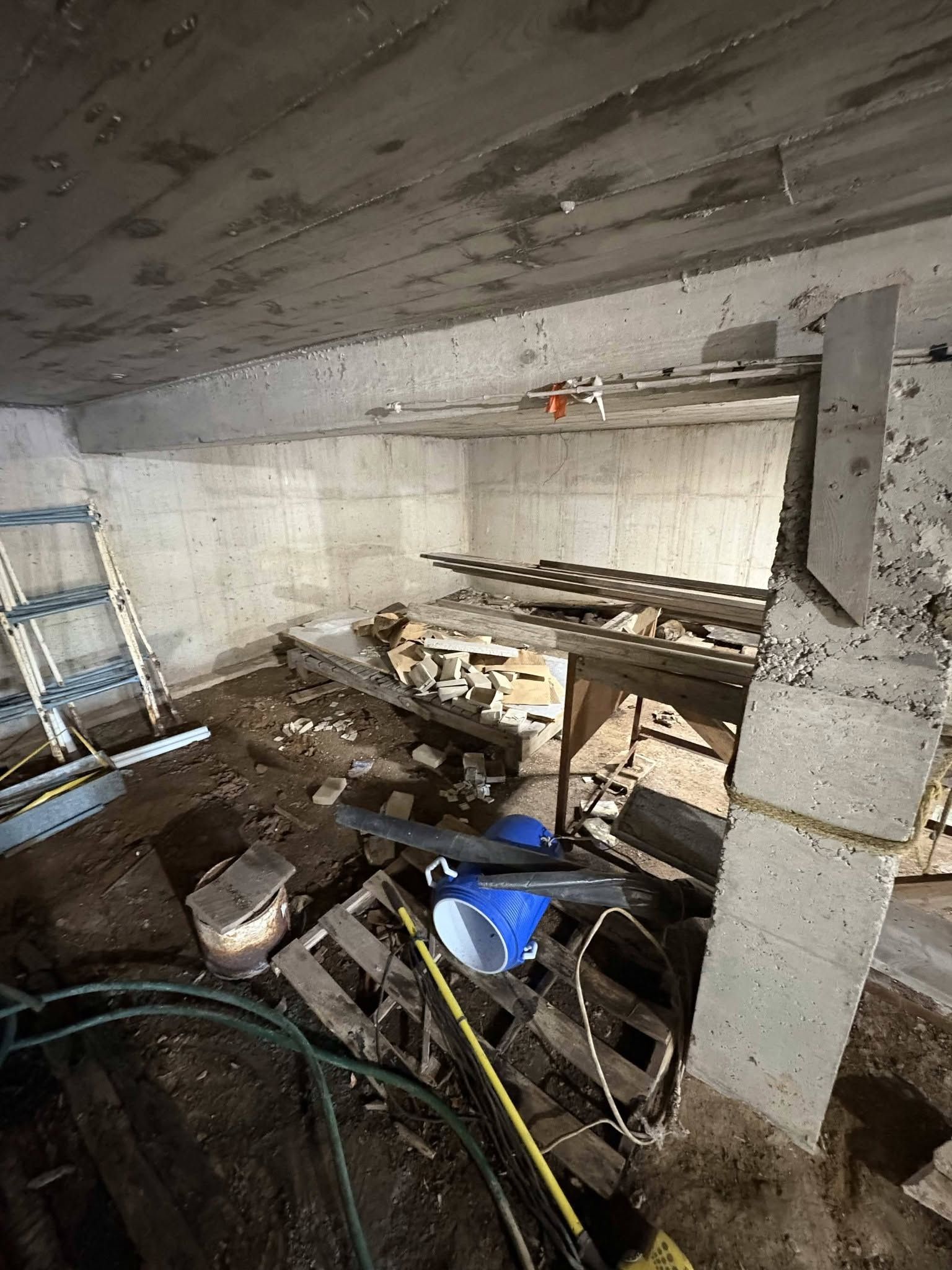 Cluttered basement with ladder, pipes, tools, and debris around a concrete support column.