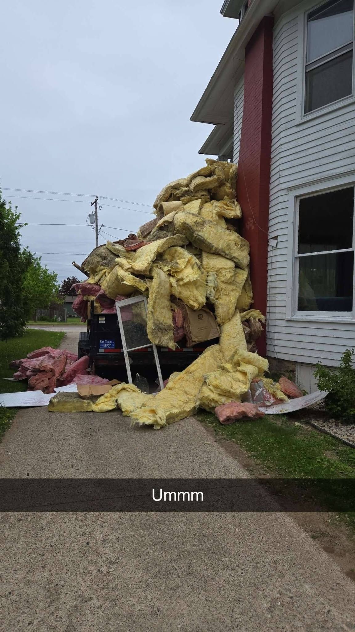 Yellow insulation debris piled against the side of a white house beside a sidewalk.