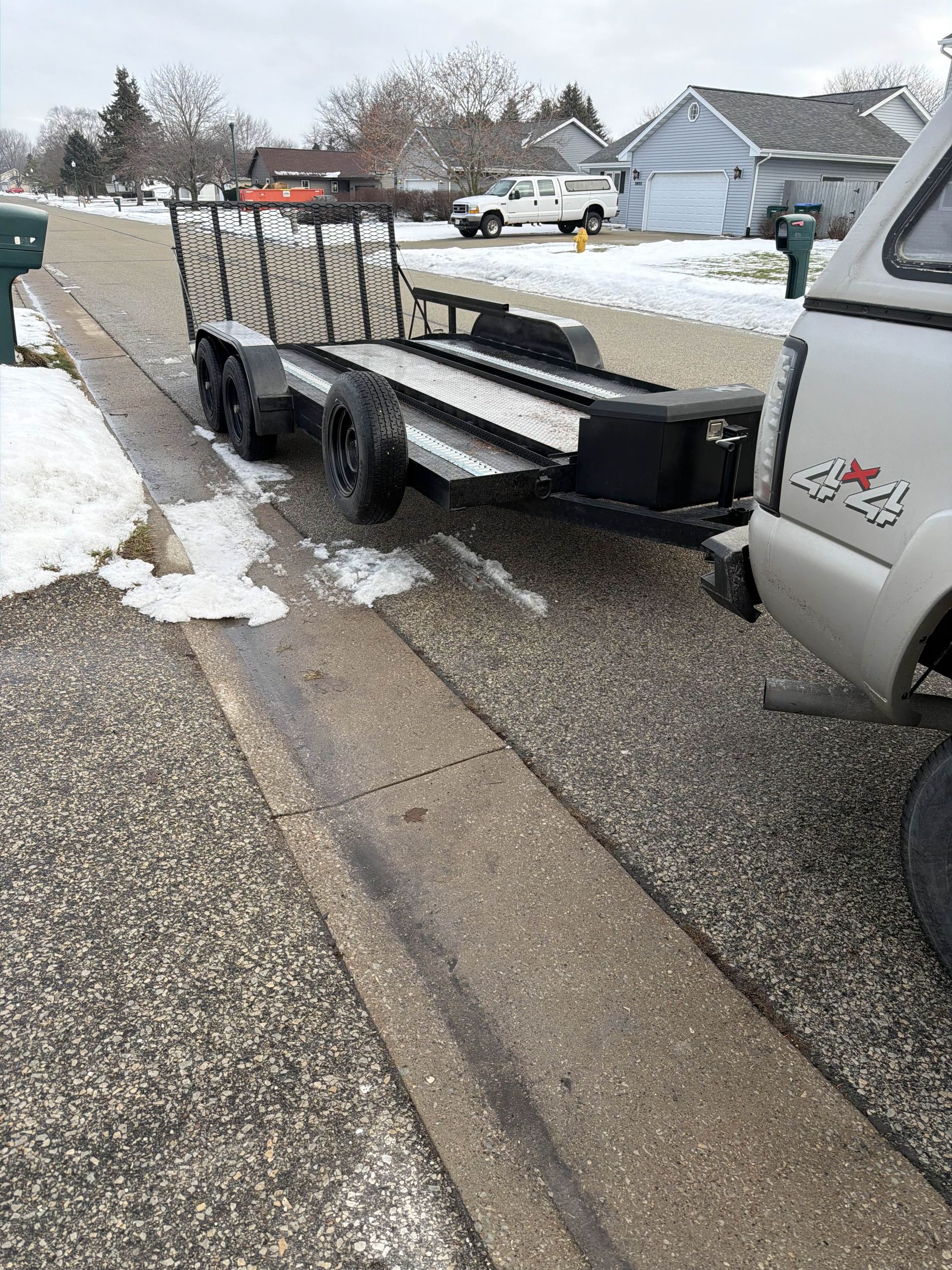 Black trailer with tools parked on snowy roadside beside a silver truck in a residential neighborhood