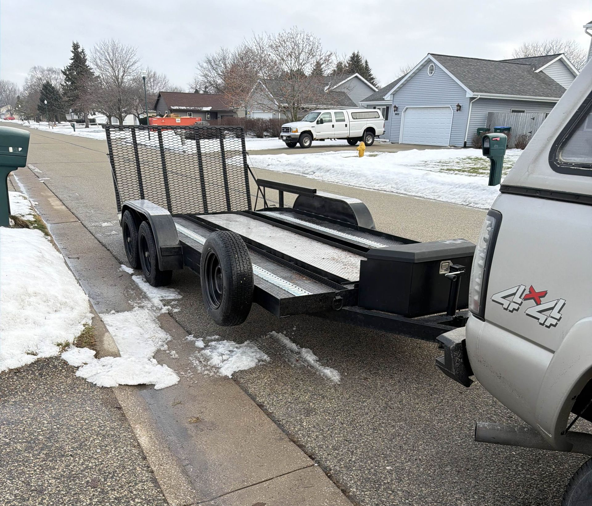 Flatbed trailer parked on a snowy suburban street beside a silver pickup truck.