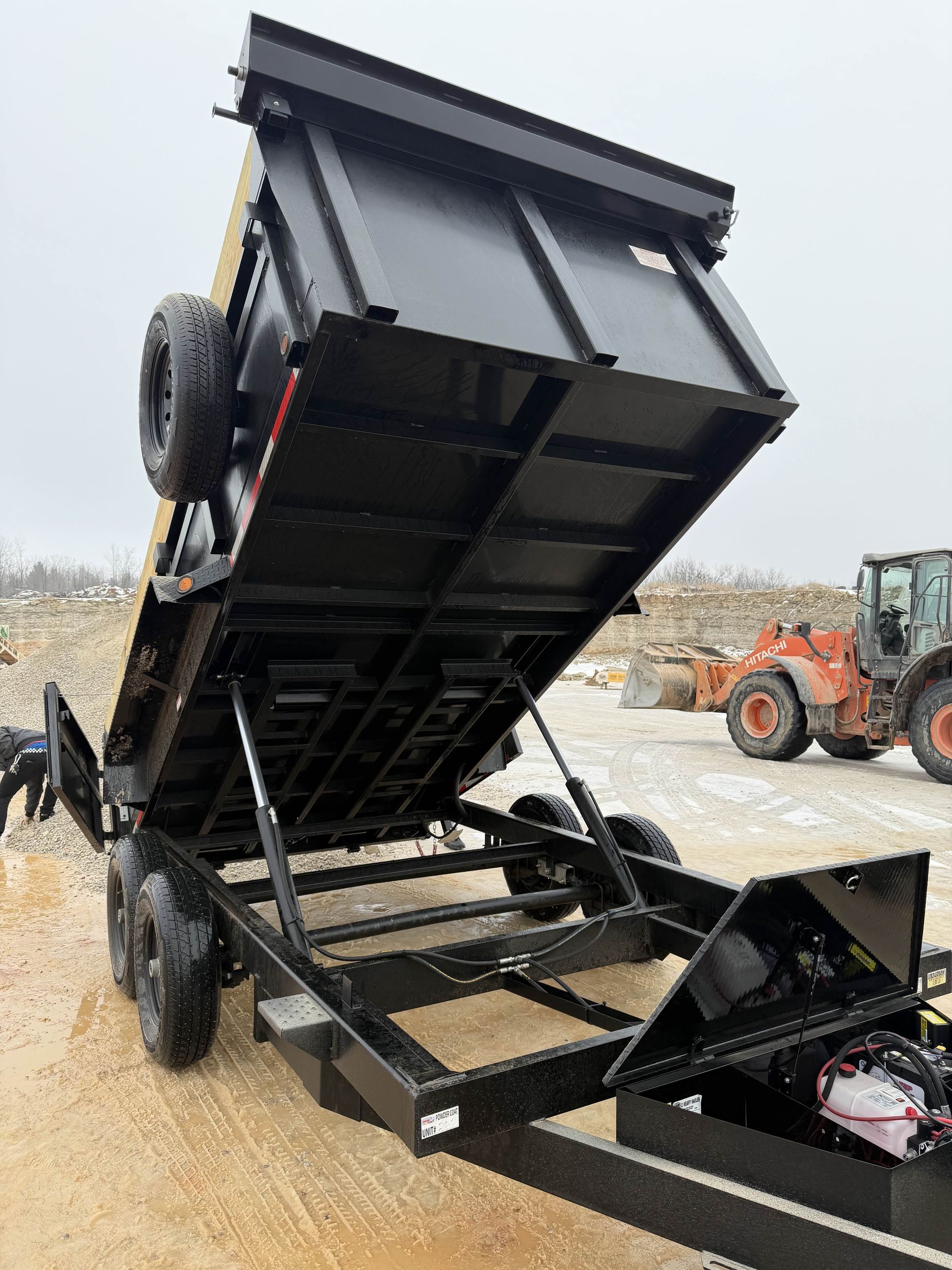 Black dump trailer tipped to unload gravel at a construction site, with a loader in the background