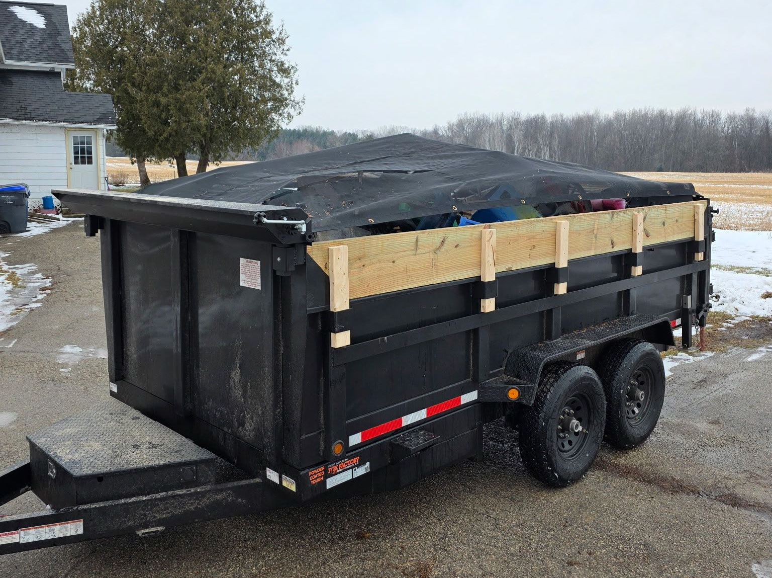 Black dump trailer filled with yard waste, parked on a snowy driveway near a house