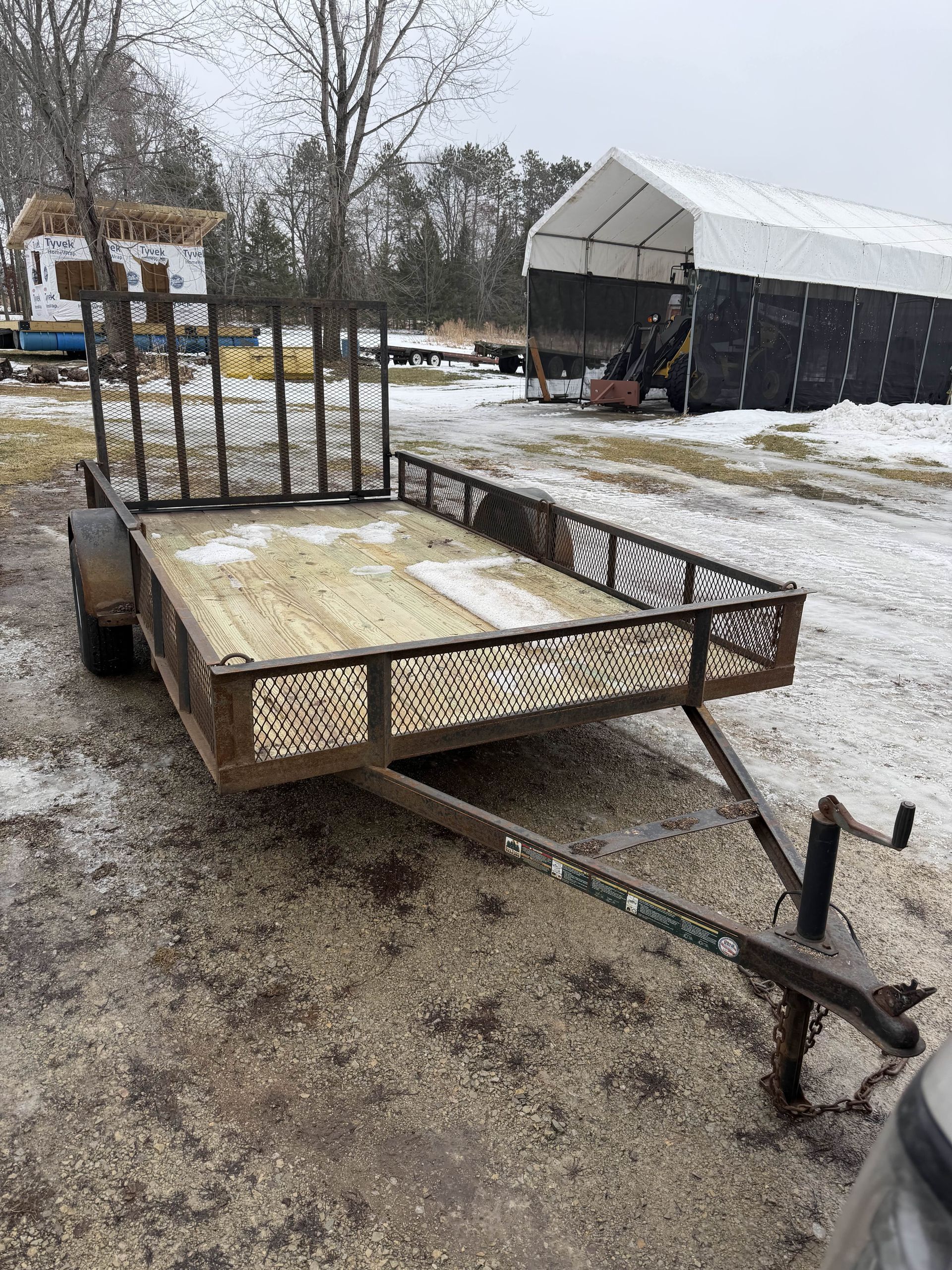 Small utility trailer with a metal gate parked on a snowy gravel lot near a barn.