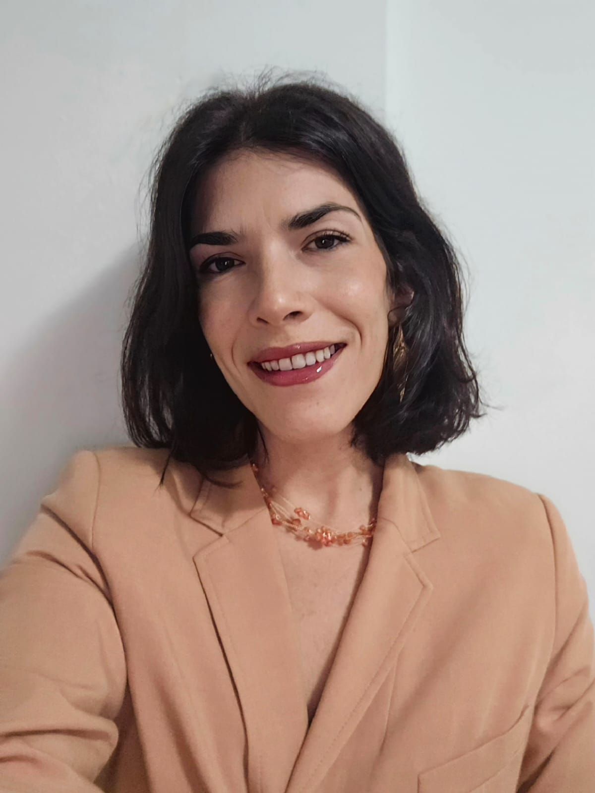 Una mujer con un blazer beige sonríe, cabello corto oscuro, collar de ámbar y fondo neutro.