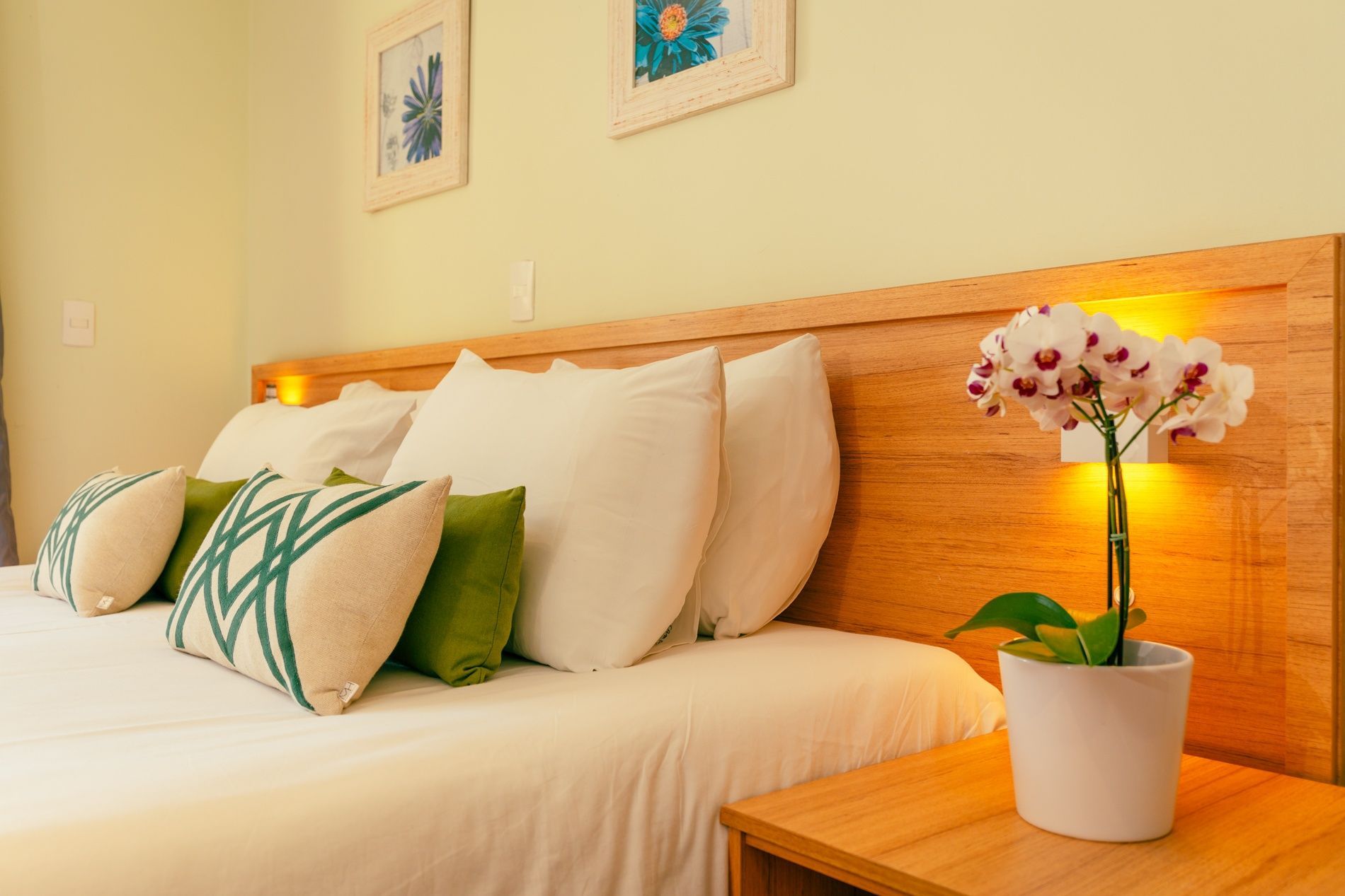 Um quarto de hotel com uma cama king-size com roupa de cama branca, cinza e laranja, e uma grande janela de vidro com vista para o banheiro.