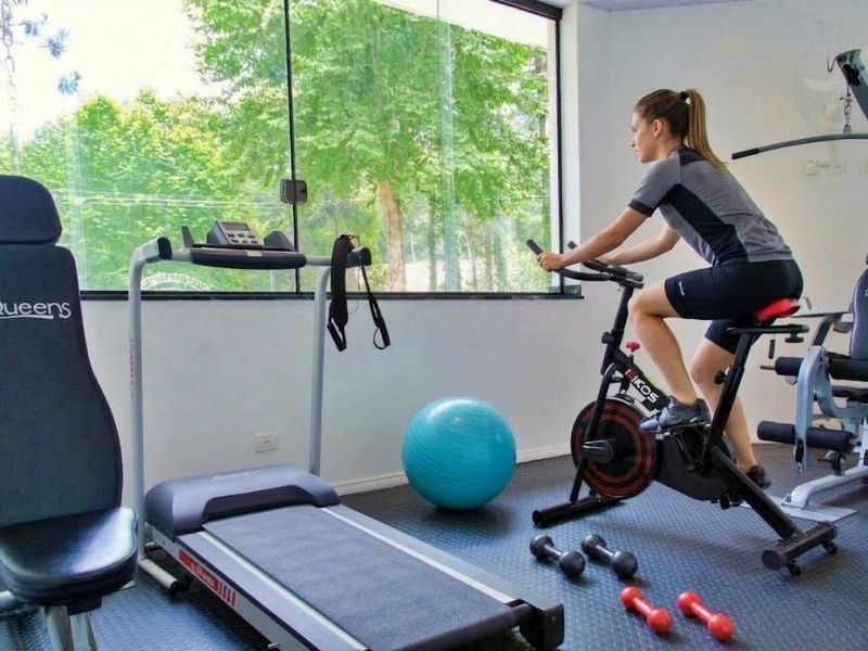 Uma academia residencial bem iluminada com esteira, elíptico, bicicleta ergométrica e um aparelho de musculação multifuncional sobre piso de madeira.