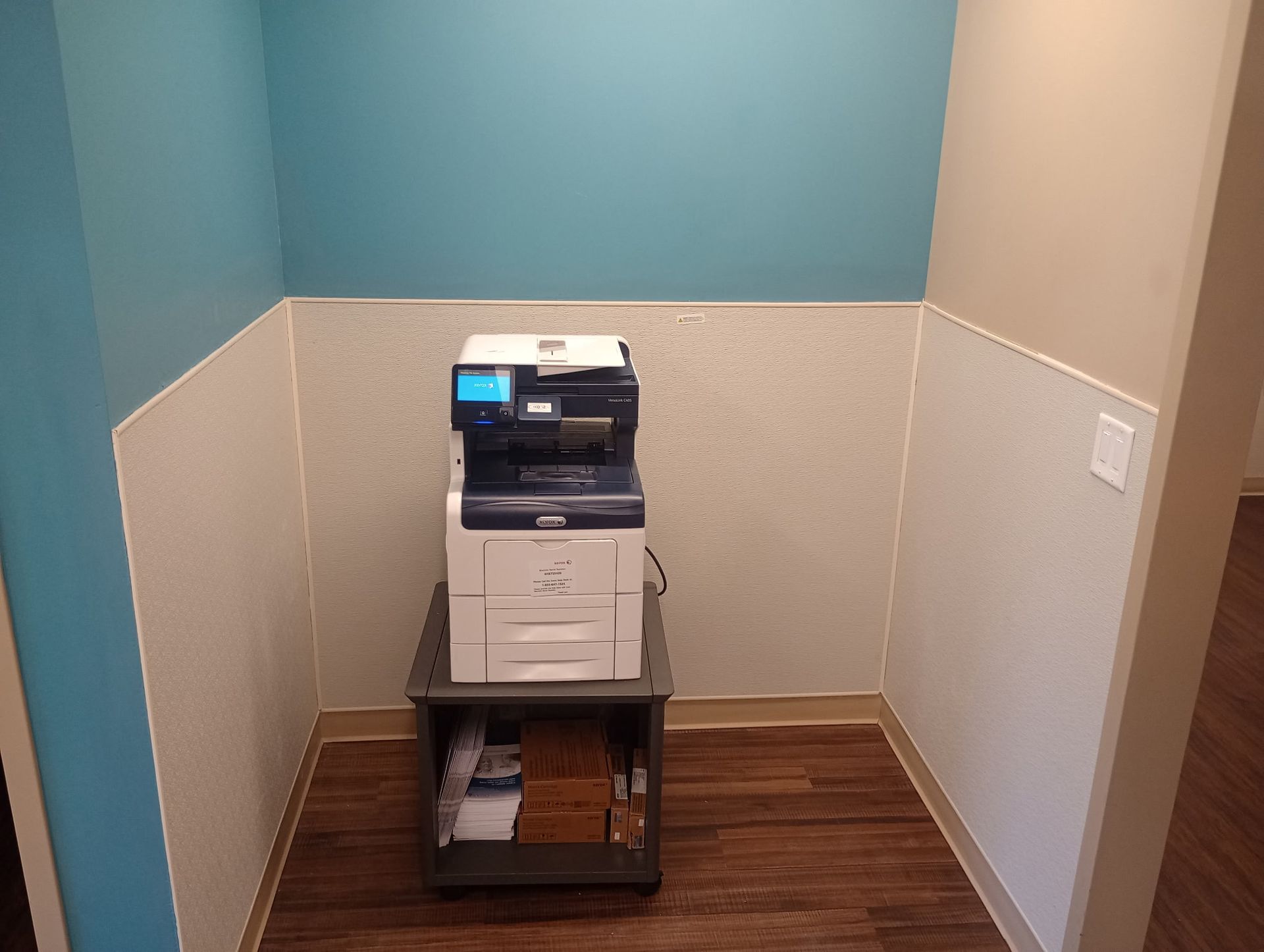 Small office copier on a rolling stand in a blue-and-beige corner nook with wood flooring.