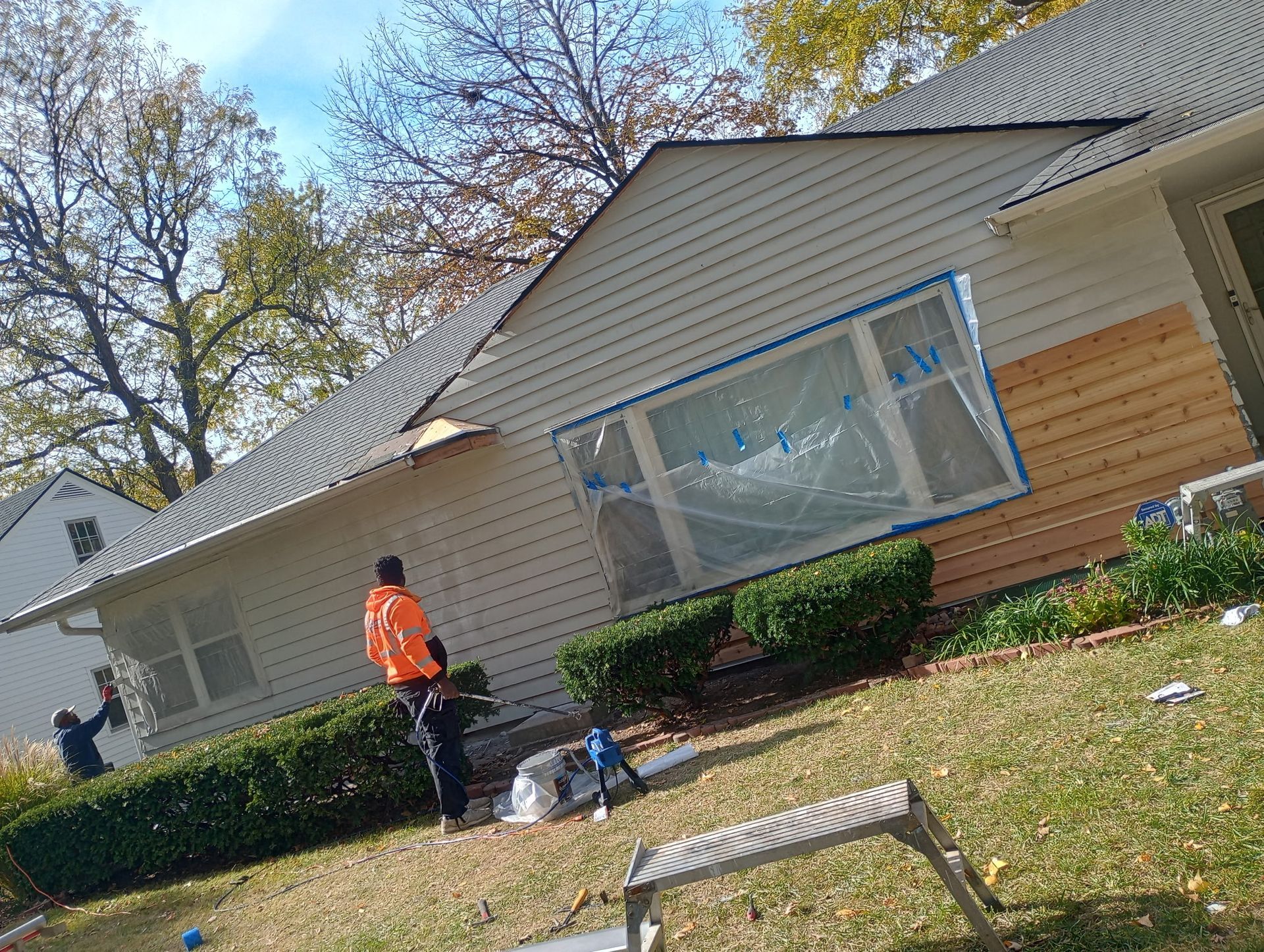 House exterior with workers wrapping a large window in blue plastic and one person in an orange safety vest
