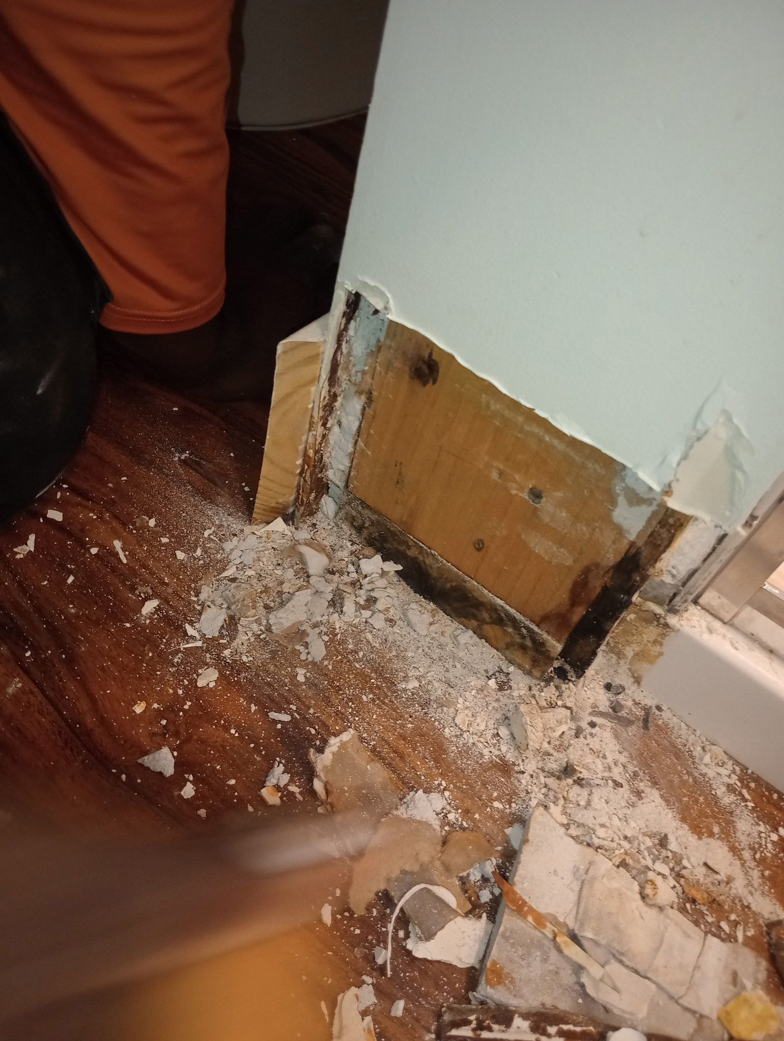 Damaged wall baseboard with exposed plaster debris on a floor beside a person’s boots