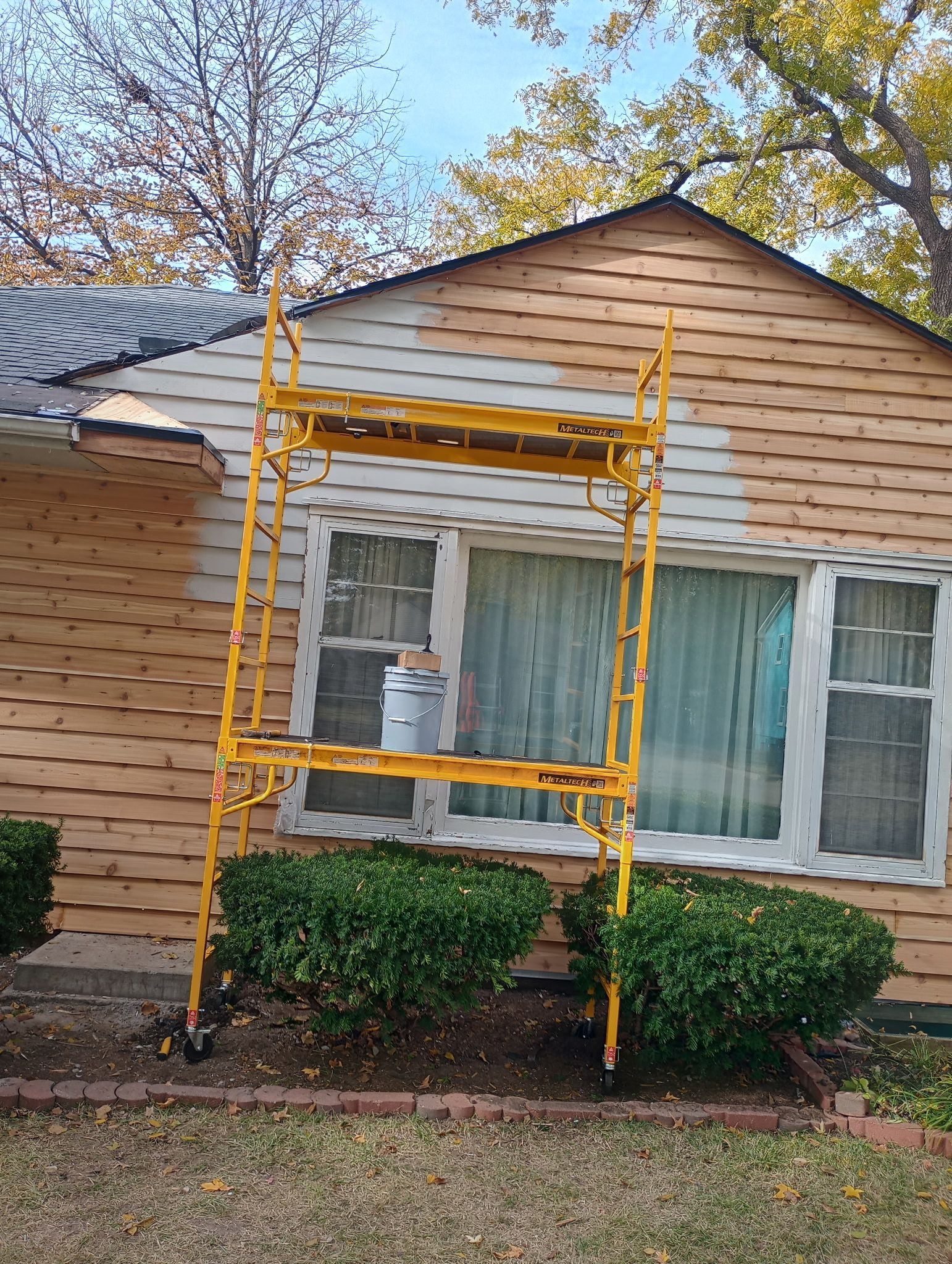 Yellow scaffolding in front of a tan house with a white-trimmed window and shrubs below.