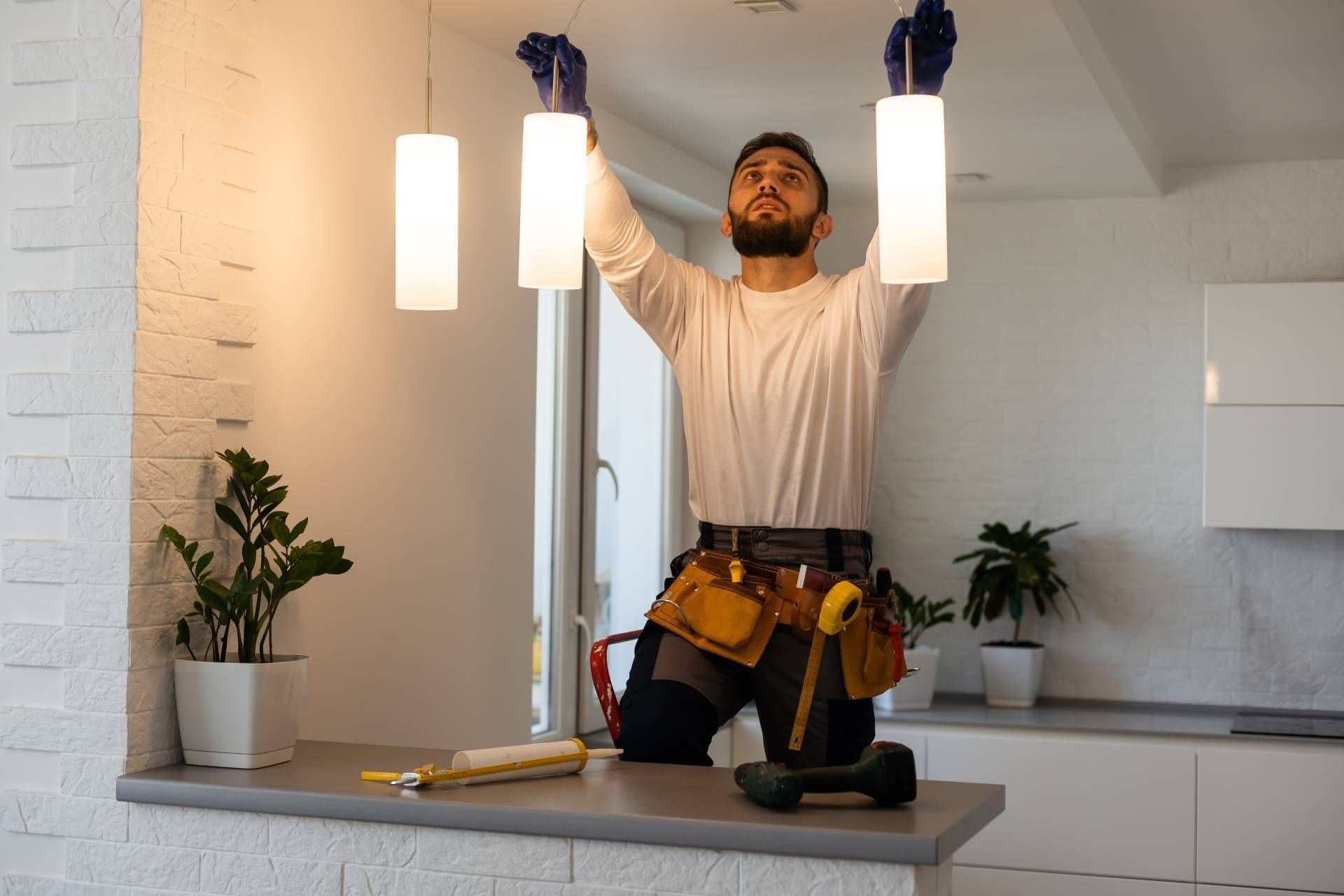 Man installing pendant lights, wearing work belt, gloves, working in a kitchen with plants.