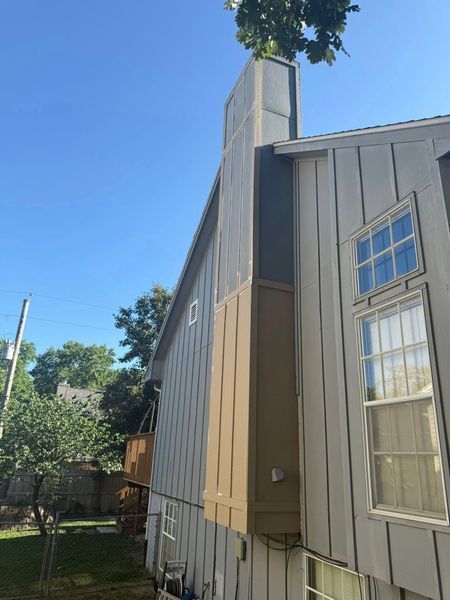 Side view of a house with light gray siding, a brown section, chimney, and windows against a blue sky.
