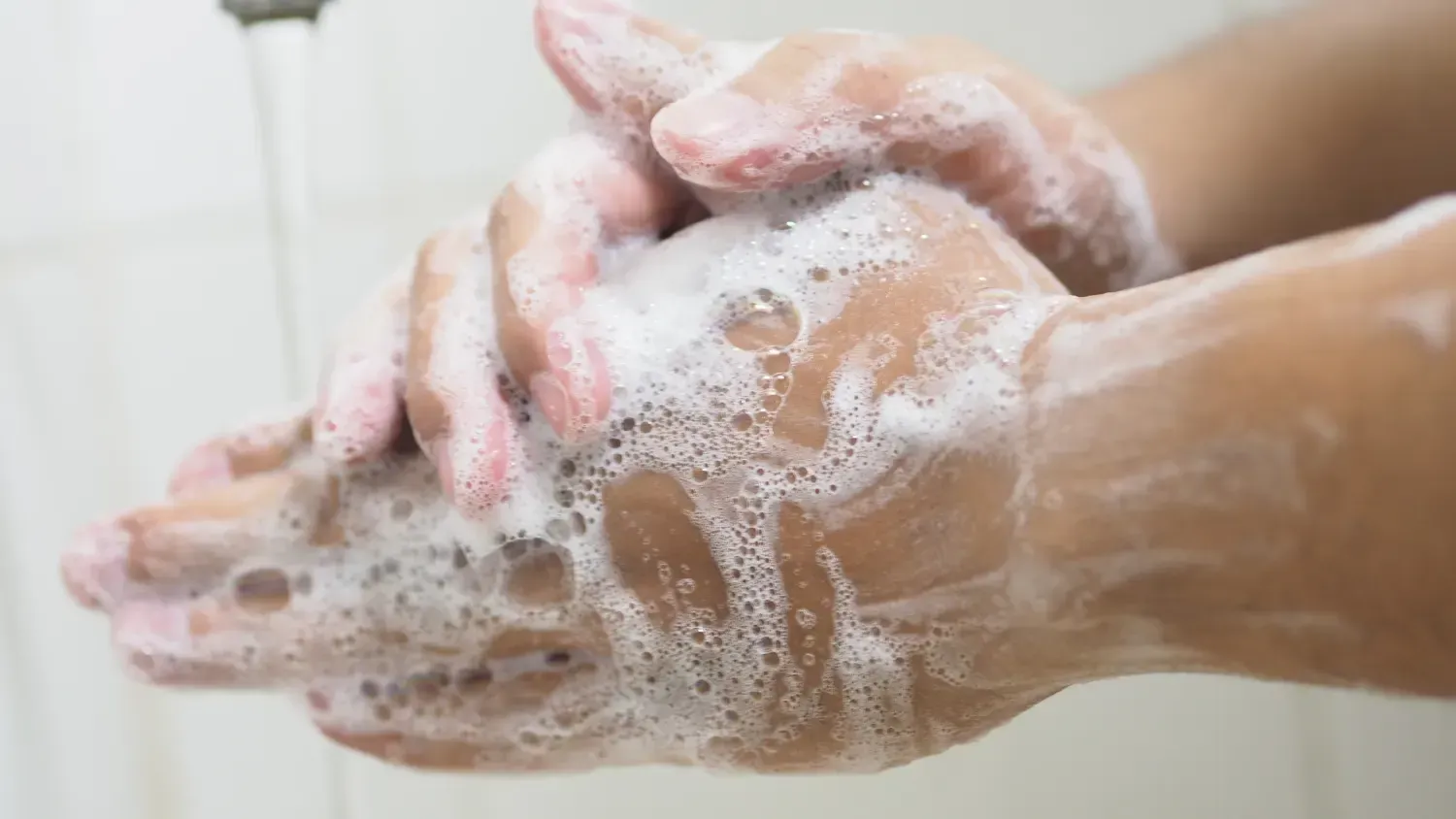 Hands covered in white, foamy soap being washed under running water.