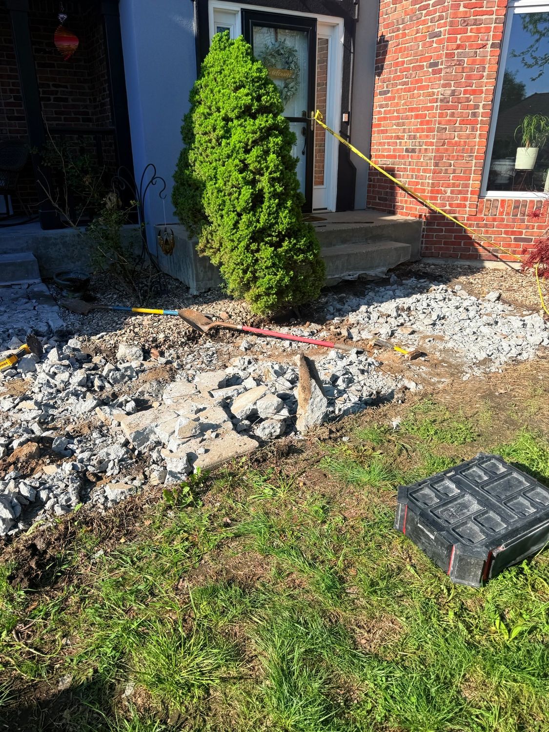 Front yard with rubble, cracked walkway, brick house entrance, and a small evergreen shrub
