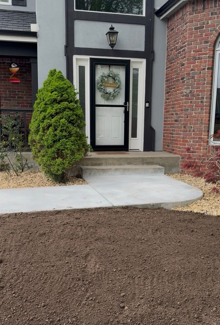 Front entrance with white door, black trim, brick wall, concrete steps, and trimmed shrub beside mulch bed
