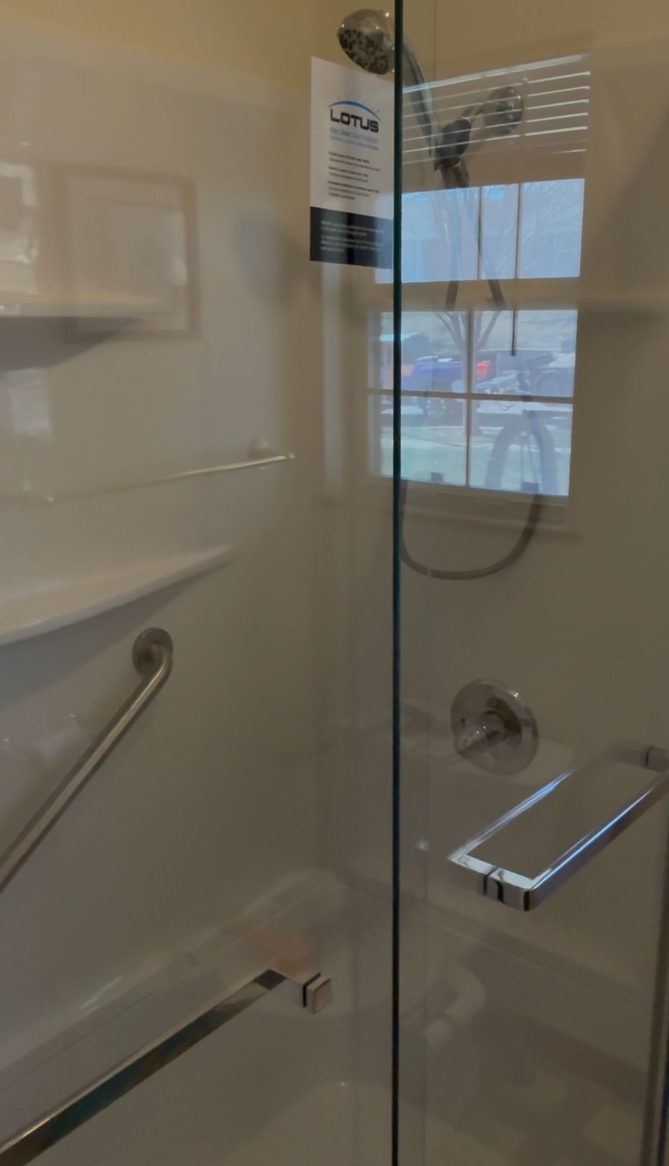 Glass shower door and chrome fixtures in a tiled bathroom, with a window reflected in the glass.