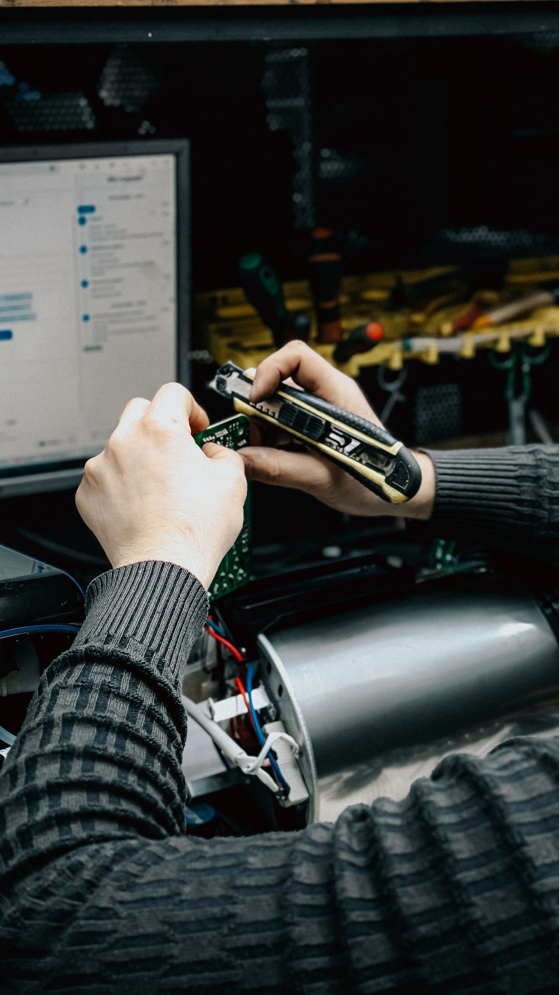 Hands working on a small circuit board in a dim workshop, with a laptop screen nearby and tools in the background.