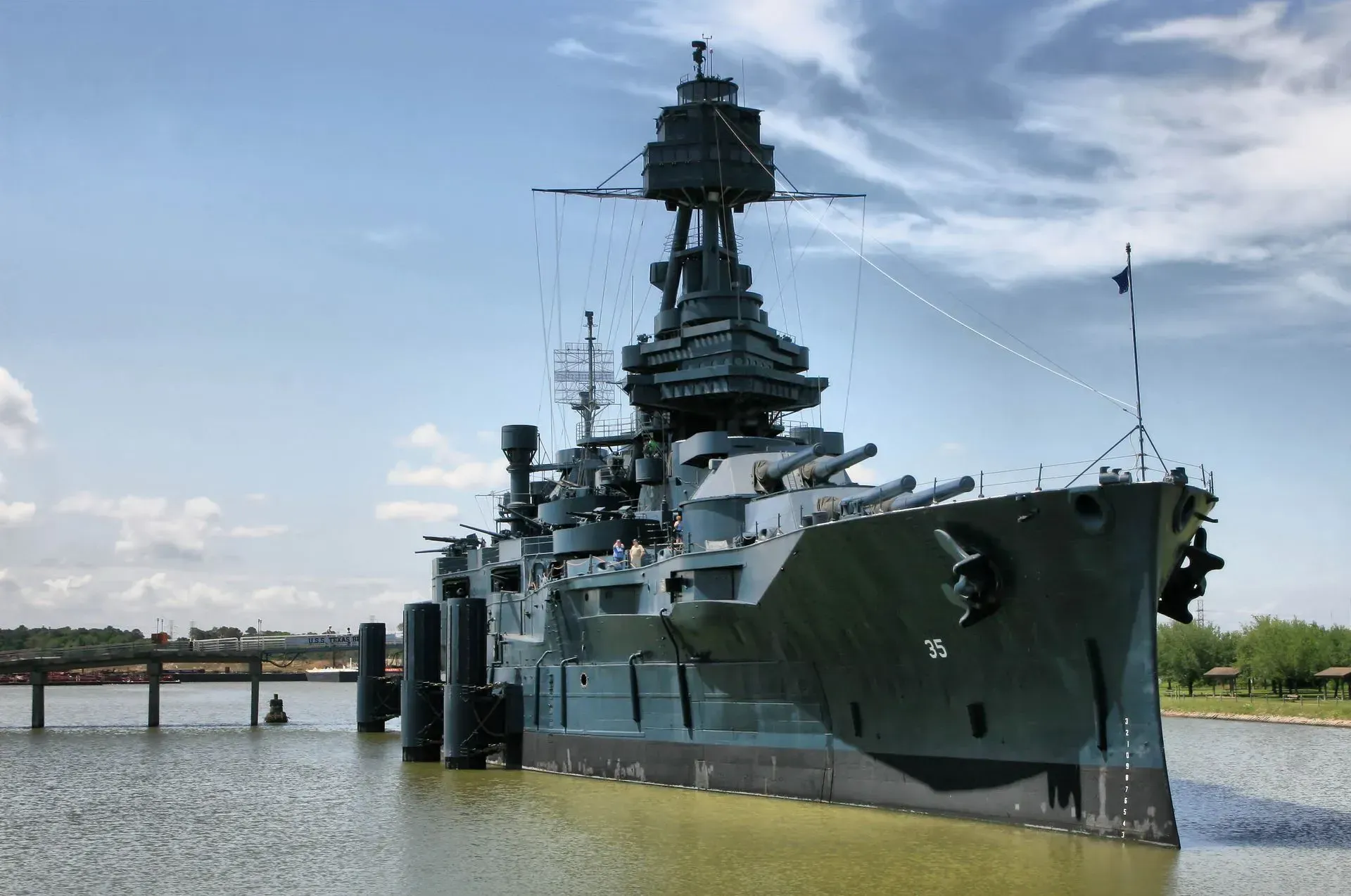 Large gray battleship docked in shallow water beside a pier under a blue sky