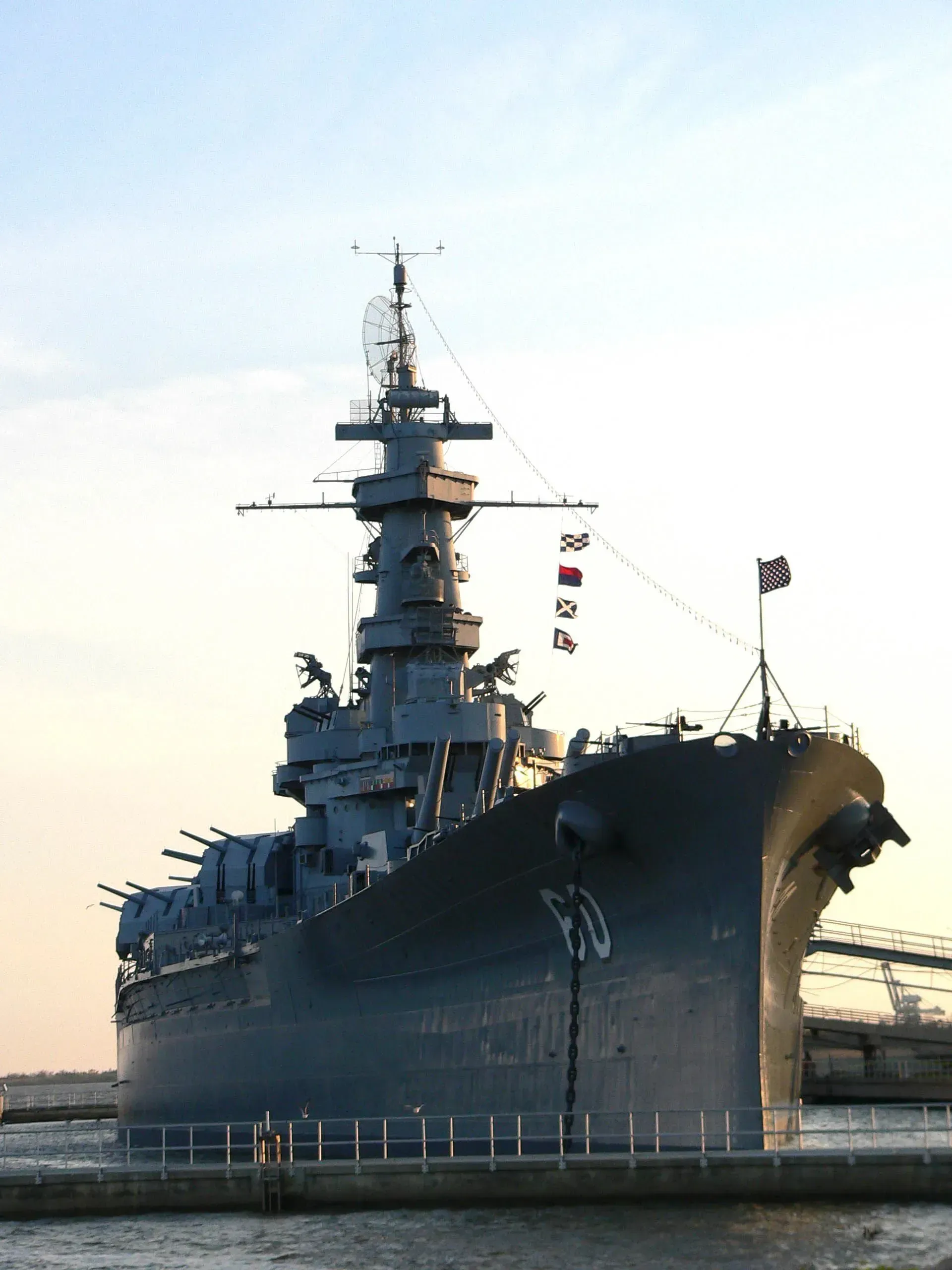 Large gray warship docked at a harbor, seen from the bow against a pale sky.