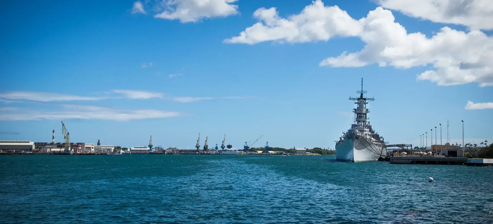 Warship at sea in a harbor with blue water, industrial shoreline, and partly cloudy sky