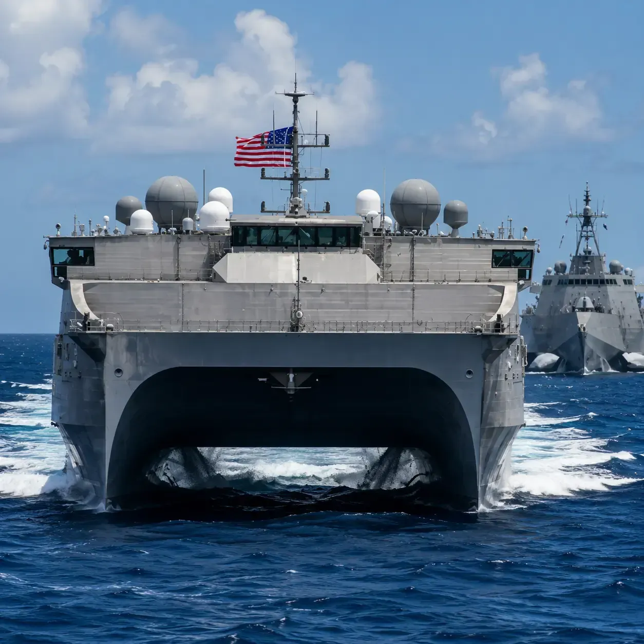 Two gray naval ships speeding across a blue ocean under cloudy sky, with an American flag on the lead ship.