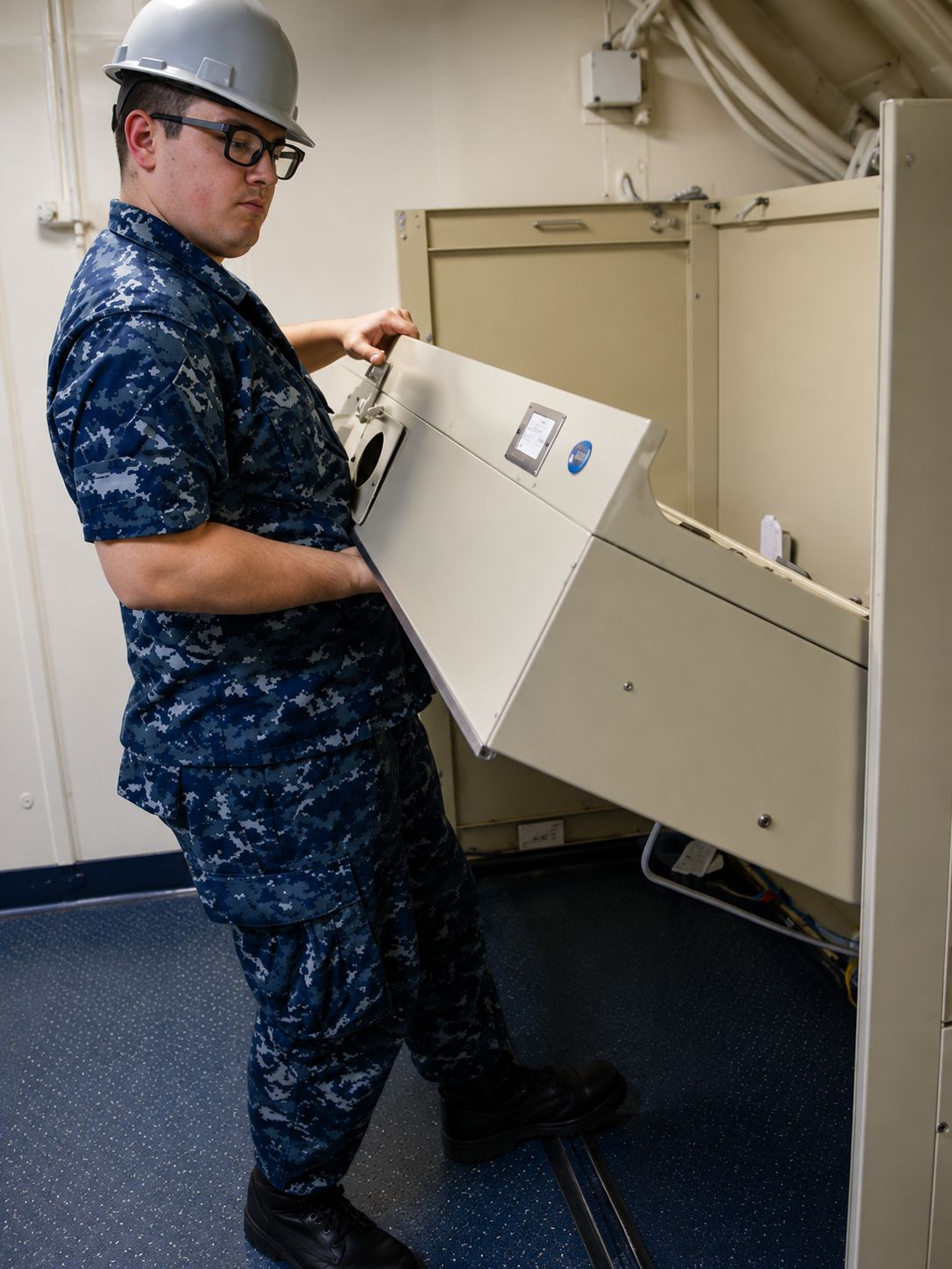Person in blue camouflage uniform lifting a beige metal panel in an industrial room