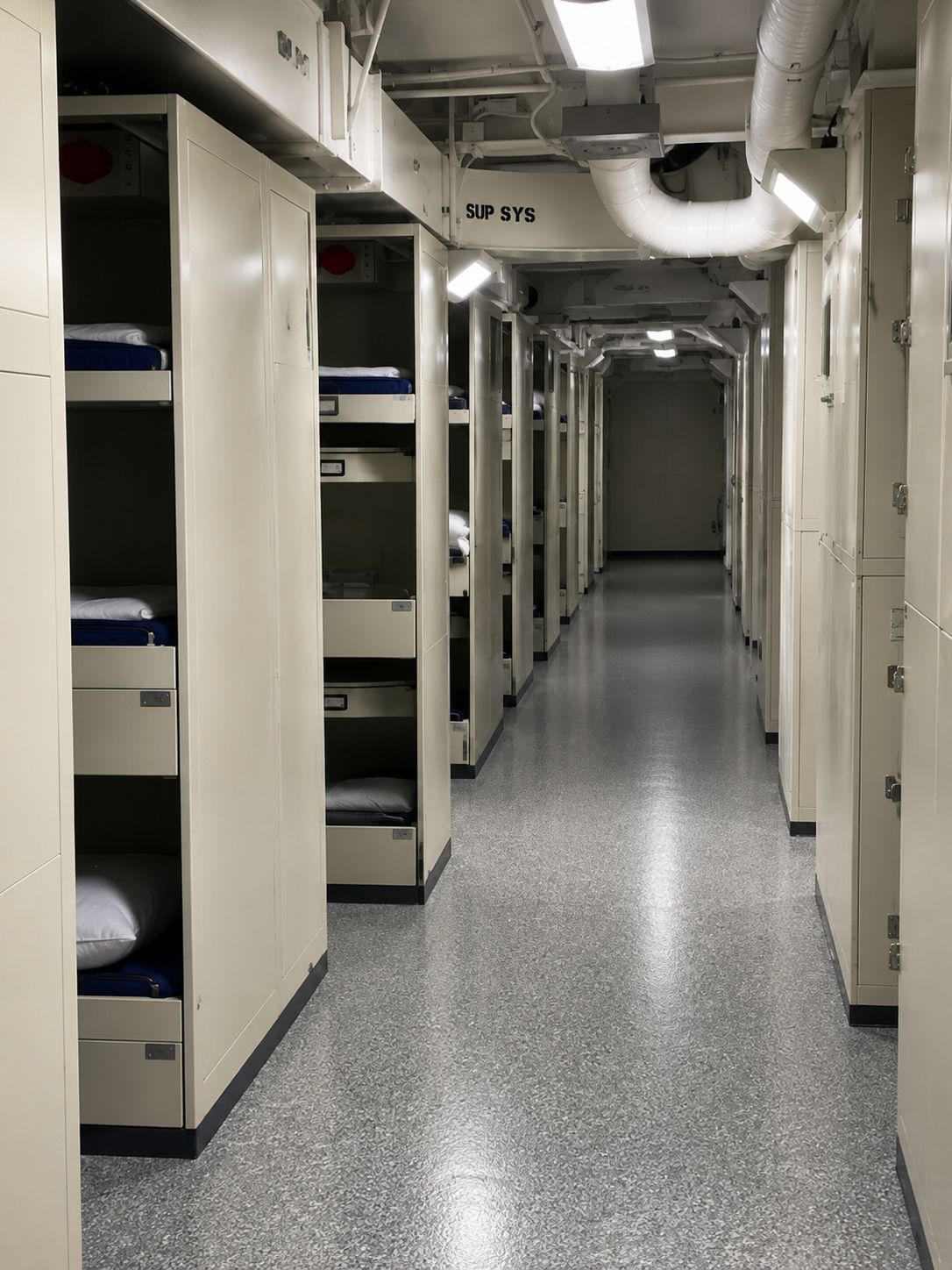 Narrow ship corridor with white bunk beds on both sides and a gray speckled floor