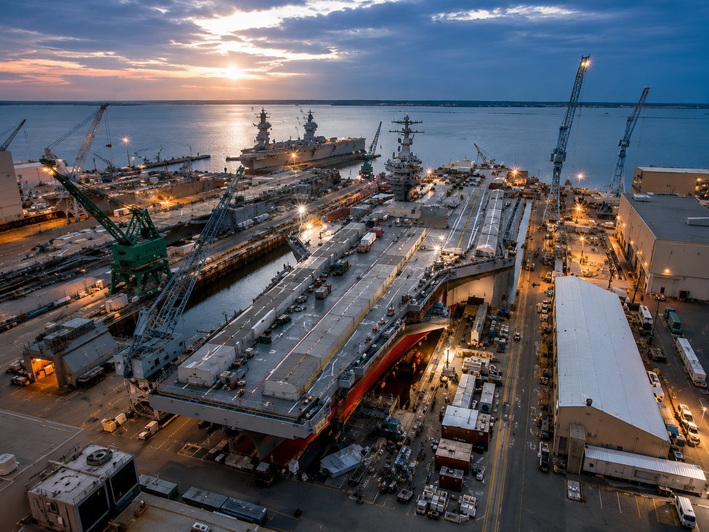 Nighttime shipyard with a large aircraft carrier under construction, cranes and lights by the water