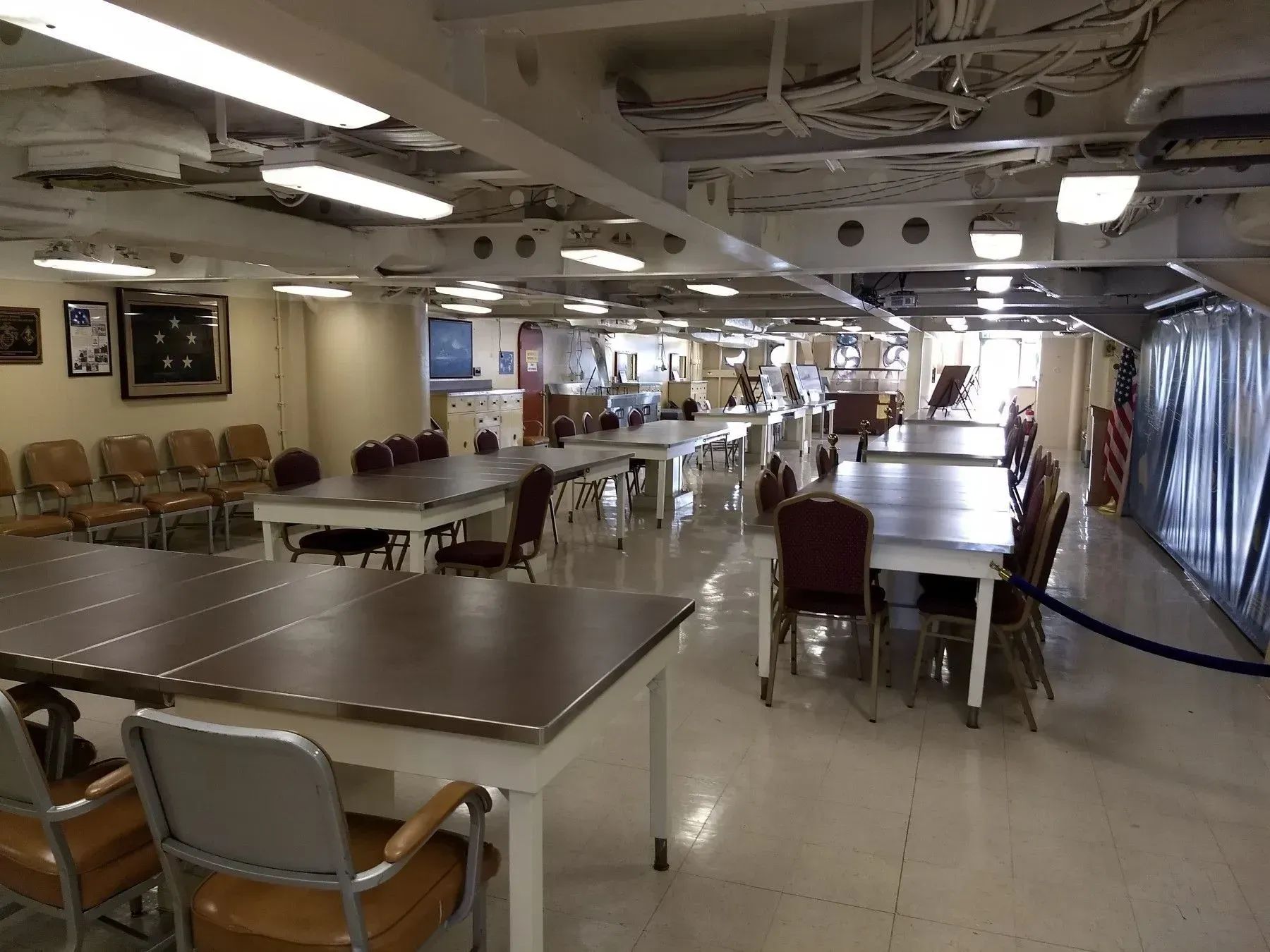 Inside a dining hall with empty tables, chairs, overhead lights, and a curtained partition on the right.