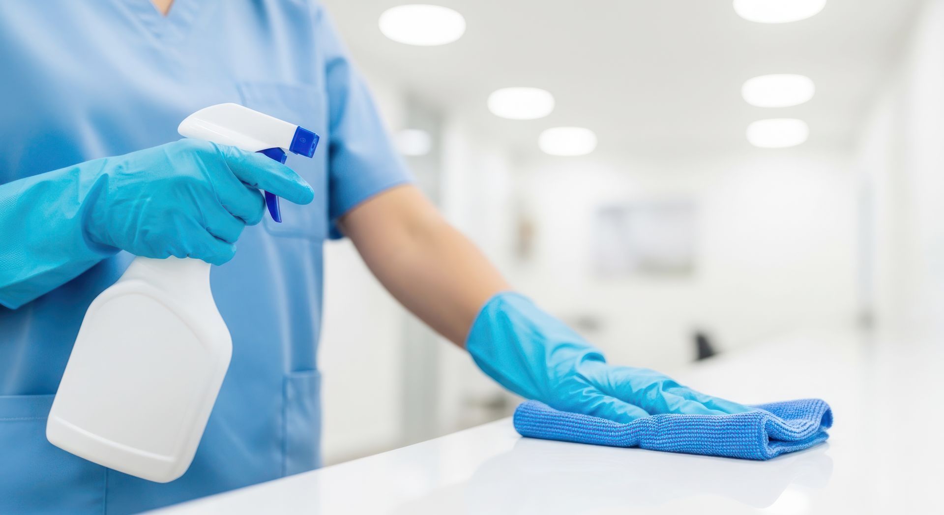 Photograph showing a person wearing blue gloves and a blue uniform cleaning a white surface with a blue cloth while holding a white spray bottle.