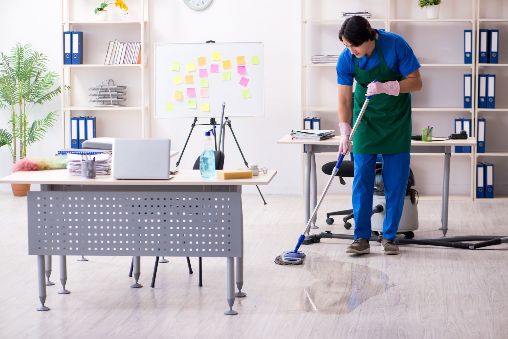 Man in blue suit, green apron vacuums an office floor. Office with desk, shelves, and a whiteboard. Man in blue suit, green apron vacuums an office floor. Office with desk, shelves, and a whiteboard.