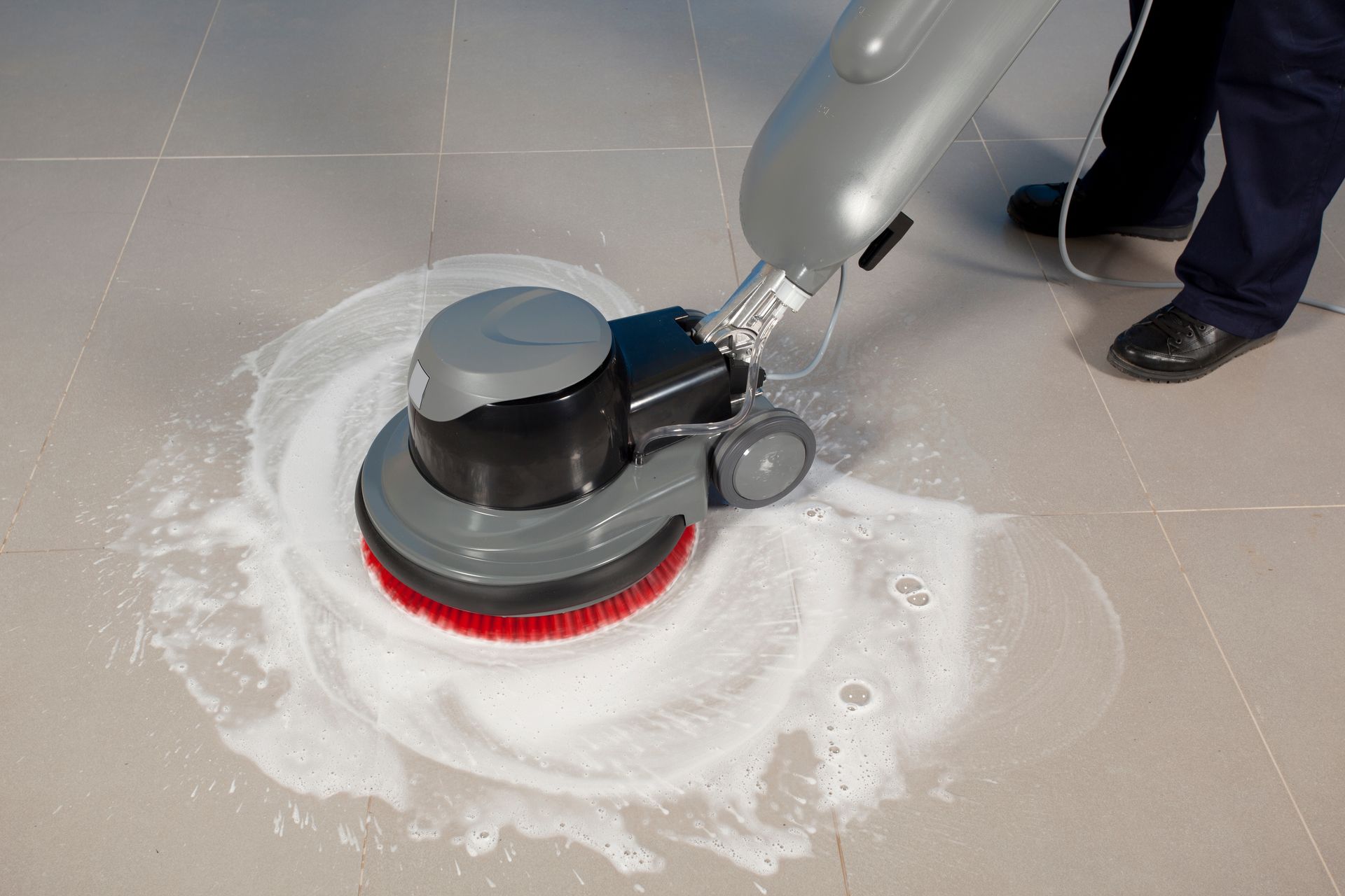 Floor scrubber cleaning a tiled floor, spreading white foam around the machine