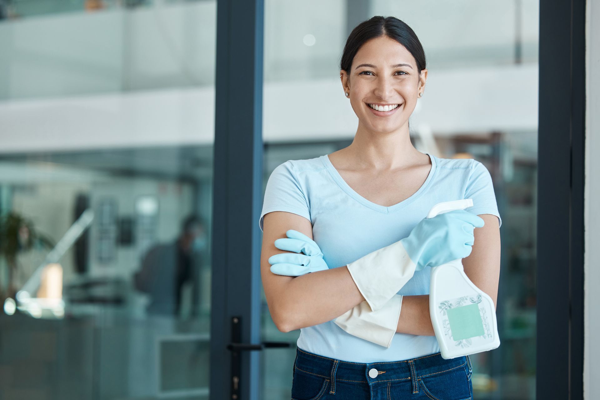 Person in blue gloves holding a spray bottle, smiling in a modern office entrance.