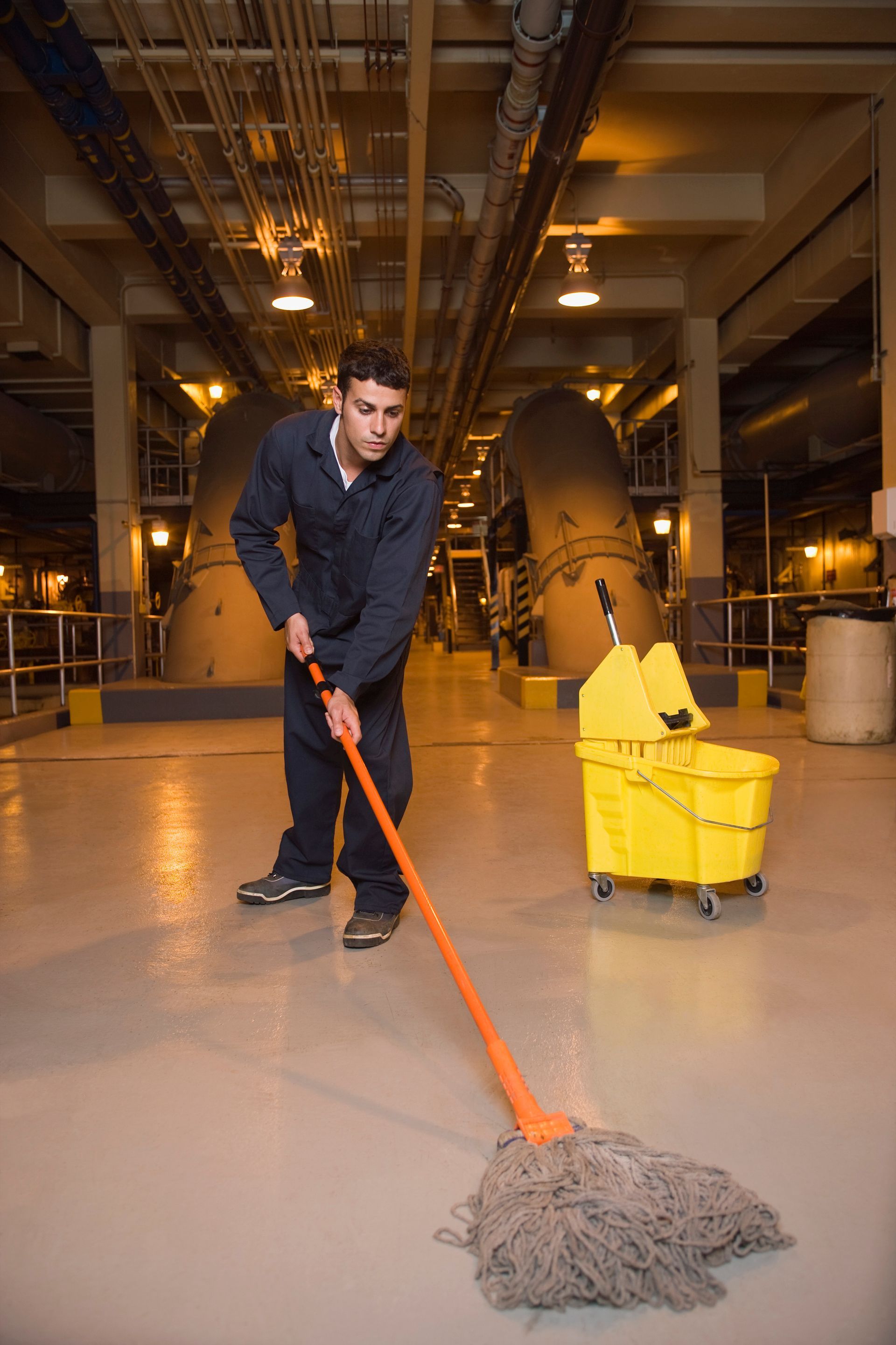 Man mops a factory floor beside a yellow mop bucket in an industrial setting