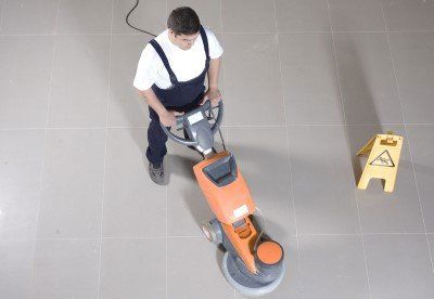 Person operating an orange floor buffer on a tiled floor beside a wet floor sign.