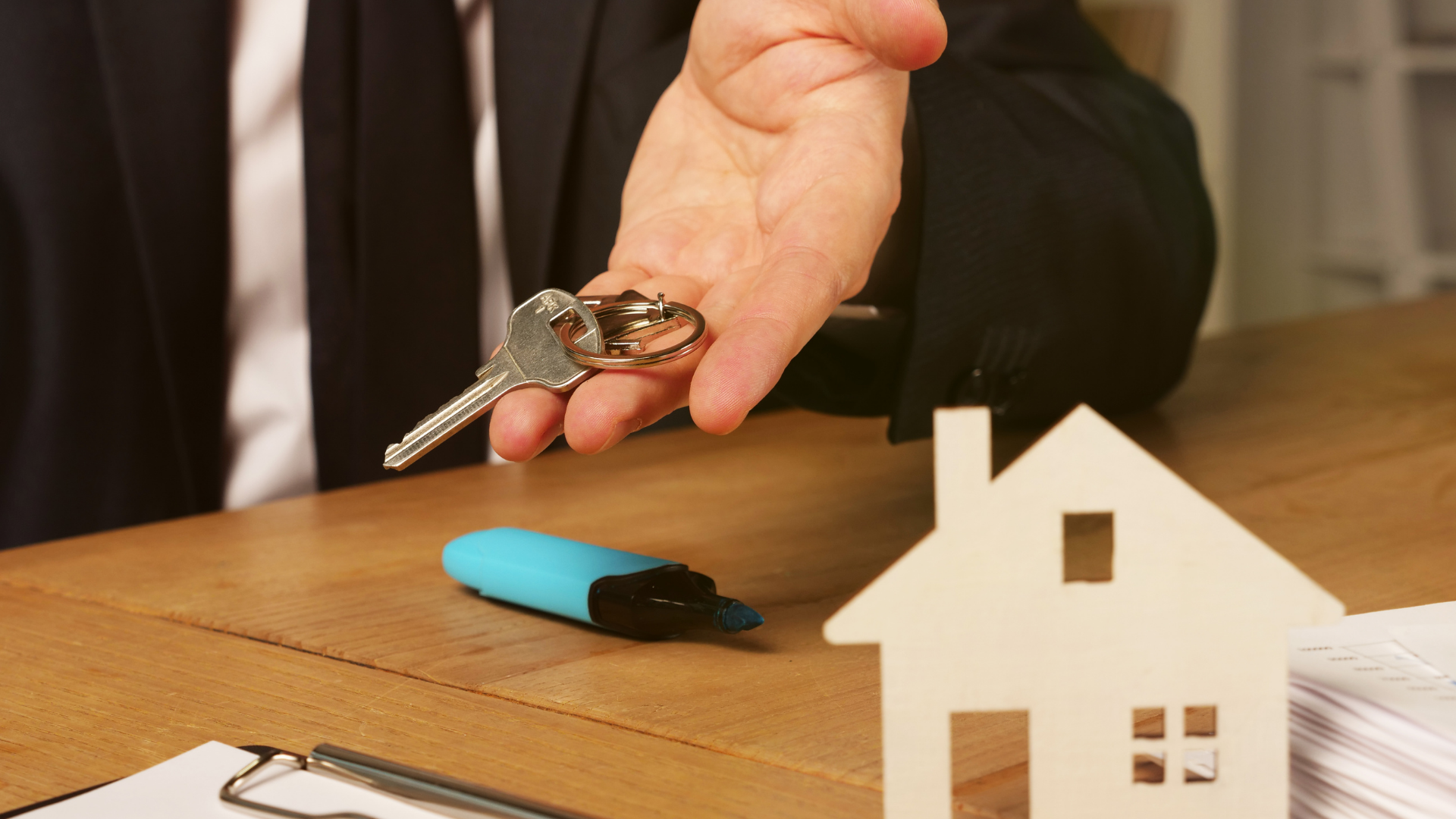 Hand holding car keys over a desk with a blue USB drive and a small white house model