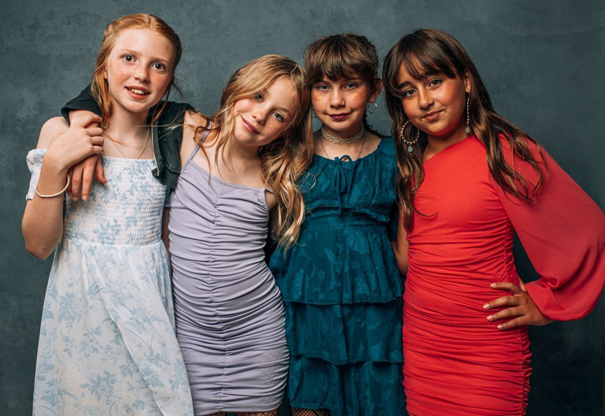 Four friends pose together, smiling in colorful dresses against a gray backdrop.