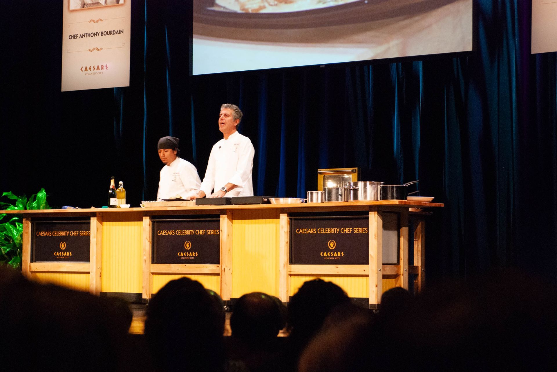 Two chefs in white uniforms working behind a counter on a stage under spotlights